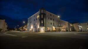 Um hotel com vários andares, entrada de canto, janelas iluminadas e área externa com mesas.