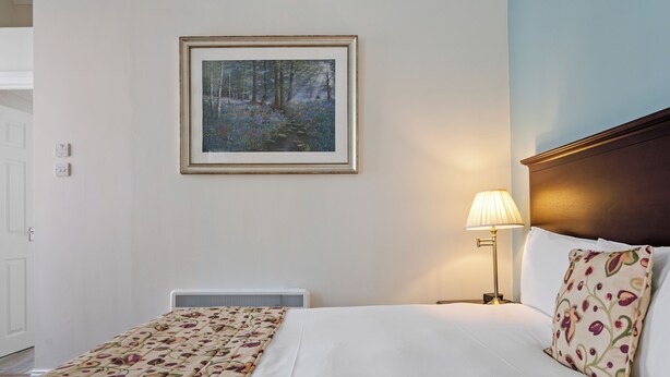 Un lit bien fait, recouvert d’une couvre-lit à motifs floraux, un tableau encadré représentant une scène de forêt accroché au mur et une lampe de chevet.