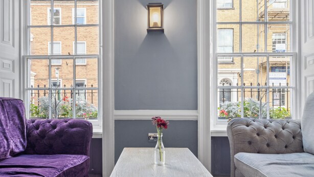 Una habitación con un sofá morado, un sofá gris y una mesa blanca con un jarrón de flores.