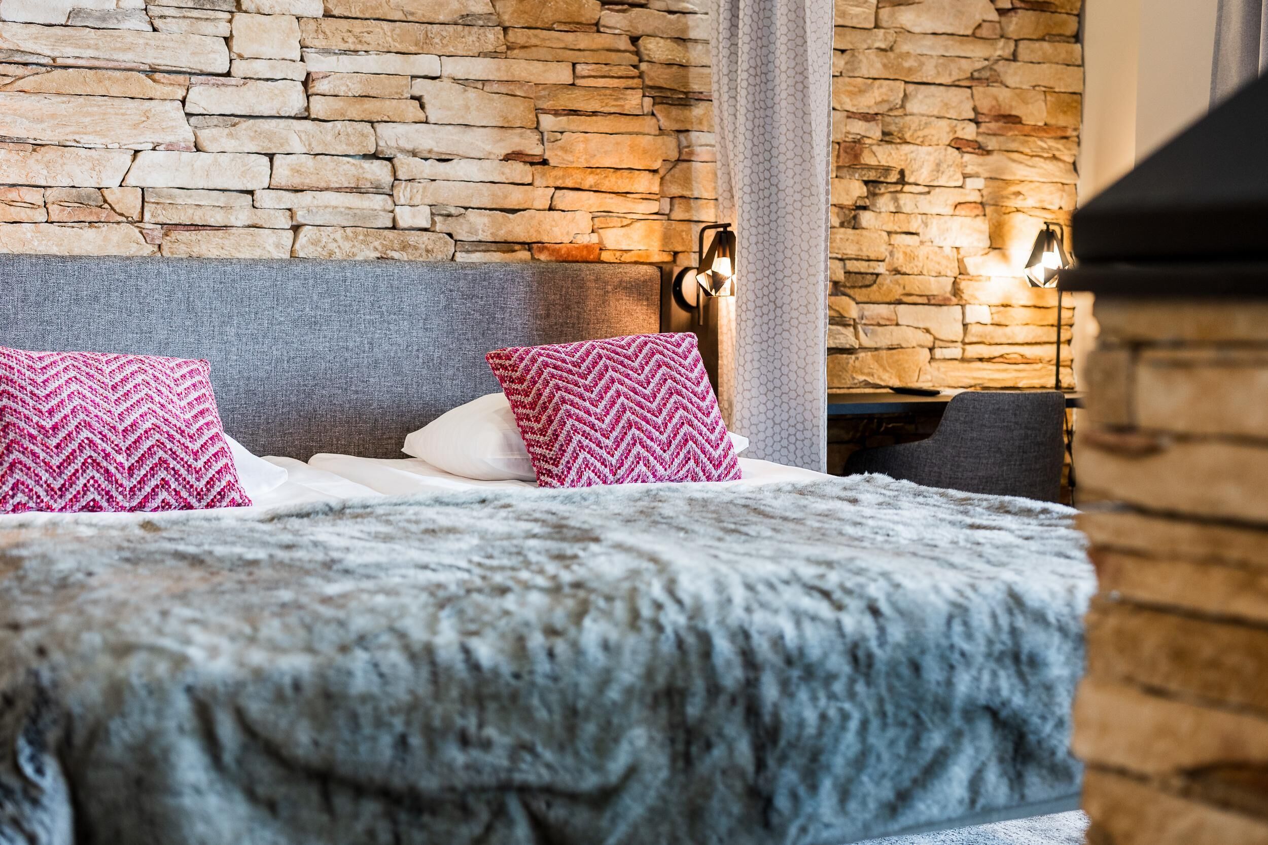 Una cama con pared de piedra de fondo, dos almohadas y una mesita de noche con lámpara.