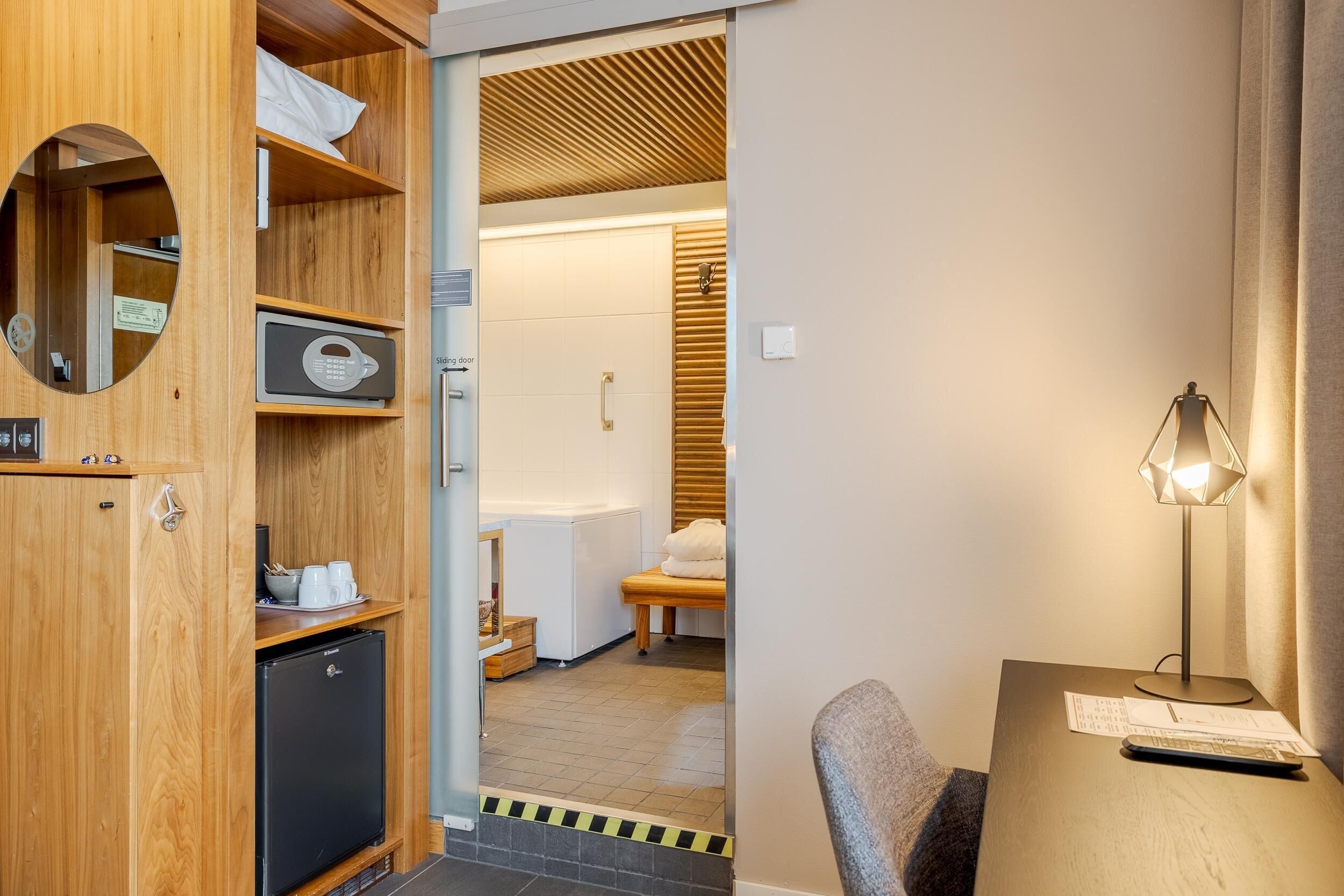 A compact hotel room with a wooden cabinet, a small refrigerator, a desk with a lamp, and a view into the bathroom.
