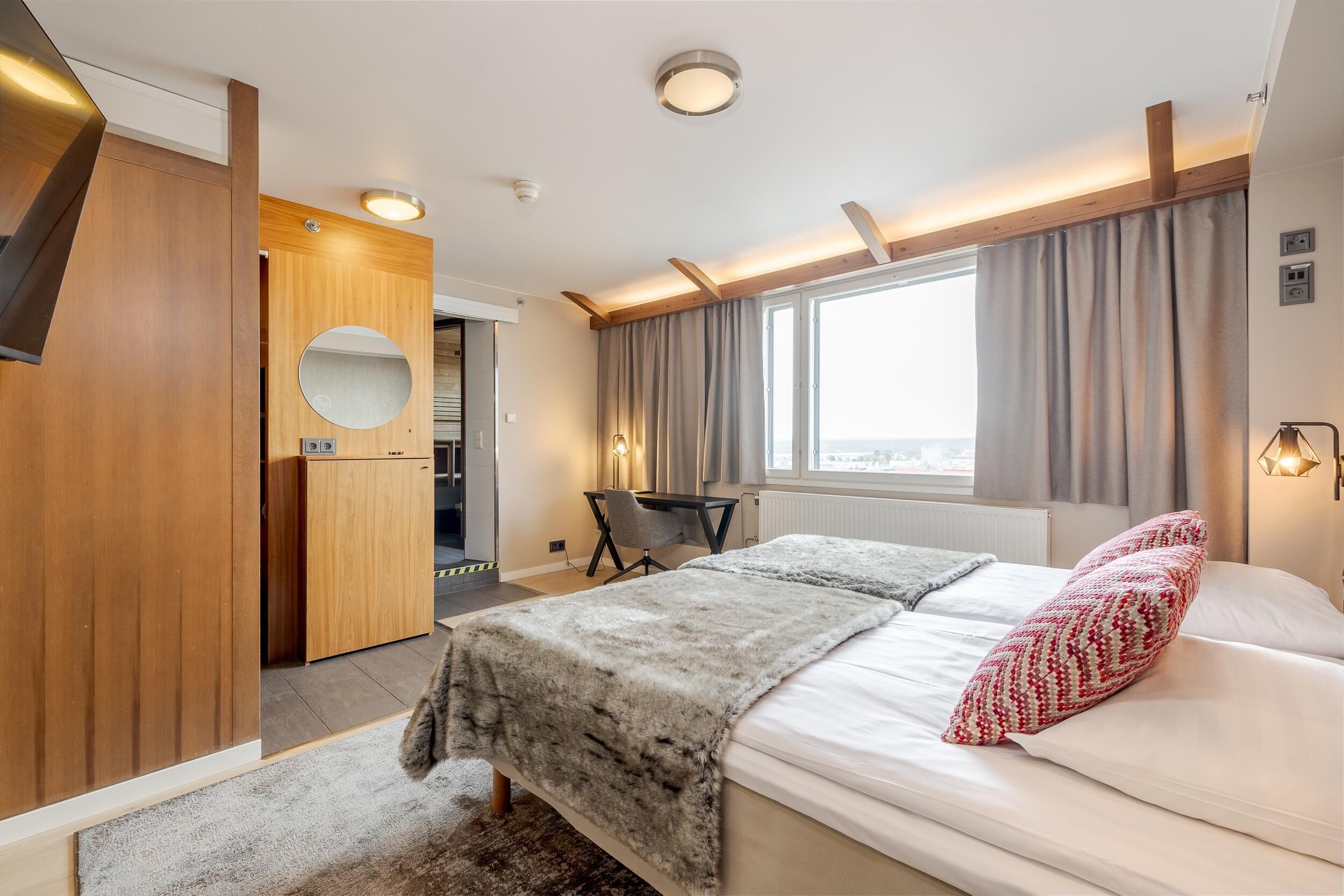 A modern hotel room with a bed, a desk, a mirror, and a view of the outdoors.