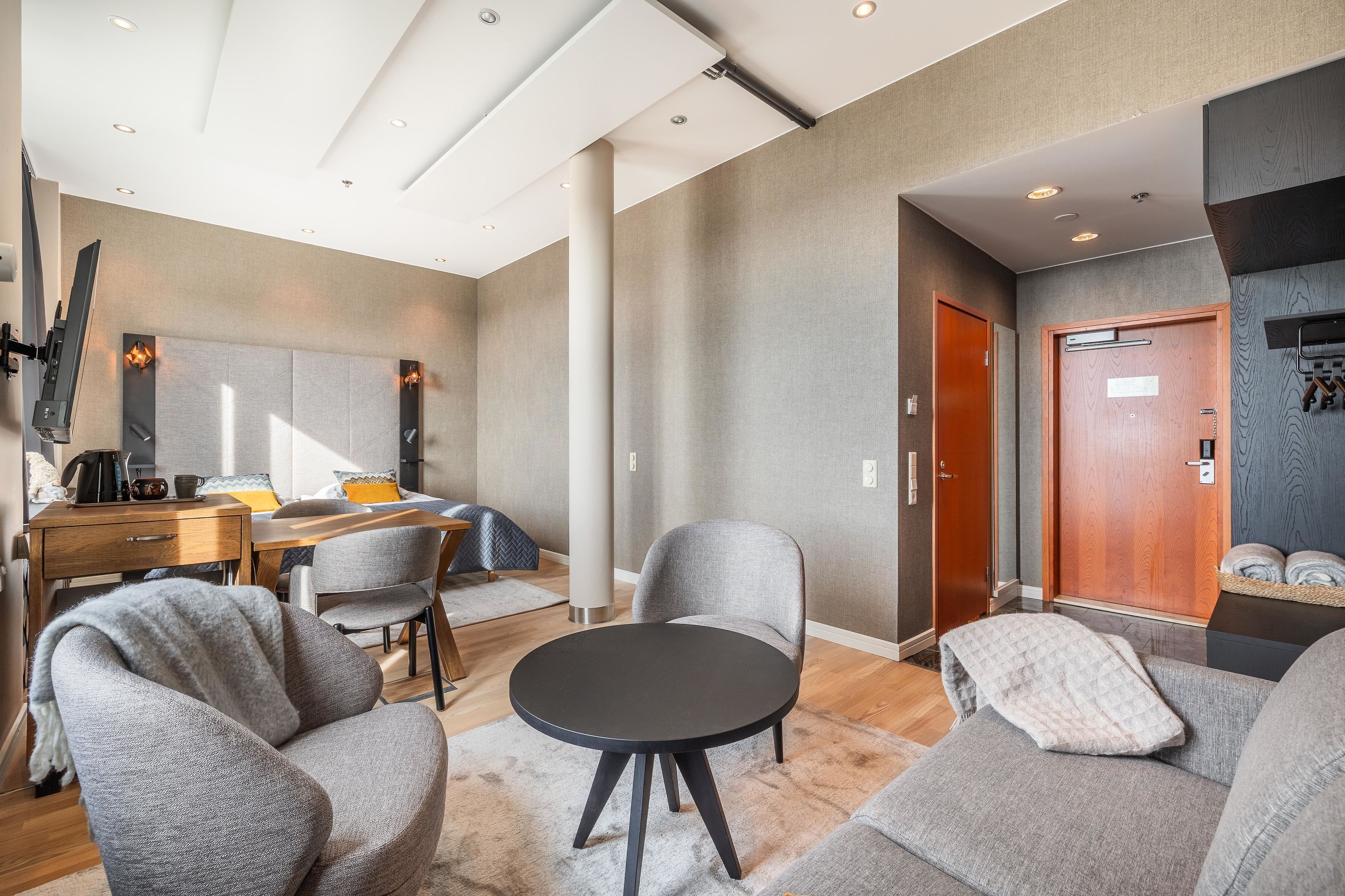 A modern hotel room with a round black table, grey sofas, a wooden desk, and a large mirror.