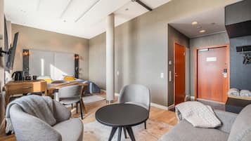 A modern hotel room with a round black table, grey sofas, a wooden desk, and a large mirror.