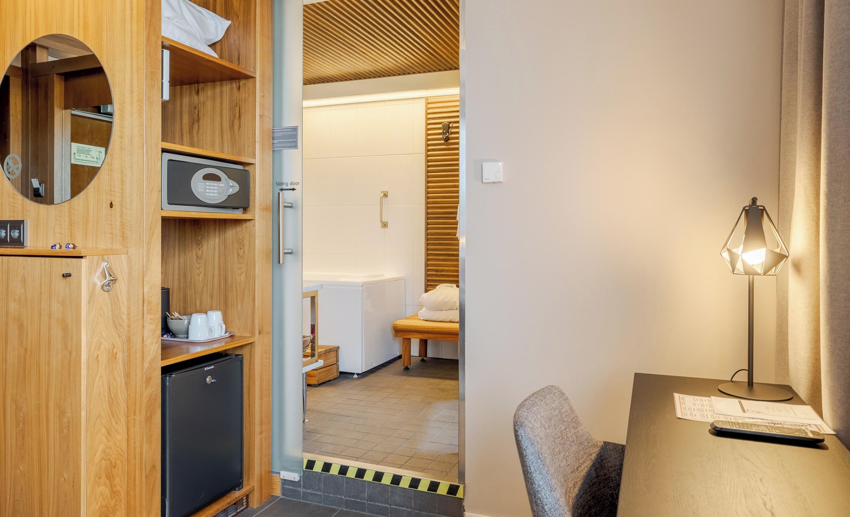 Une chambre d’hôtel avec une armoire en bois, un bureau avec une lampe et une porte coulissante donnant accès à la salle de bain.