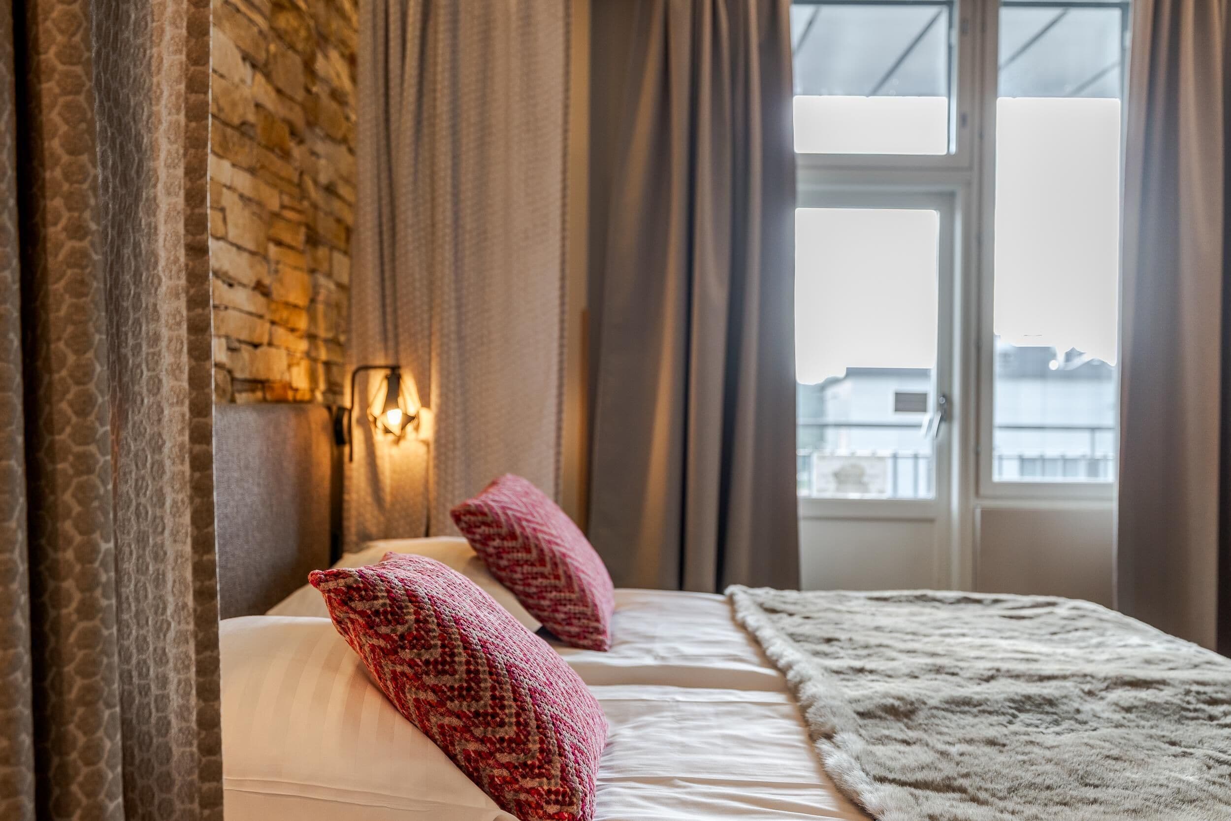 Ein Hotelzimmer mit Backsteinwand, einem Bett mit weißer Bettwäsche und zwei roten Kissen, einer flauschigen weißen Decke und einem Fenster mit Vorhängen.