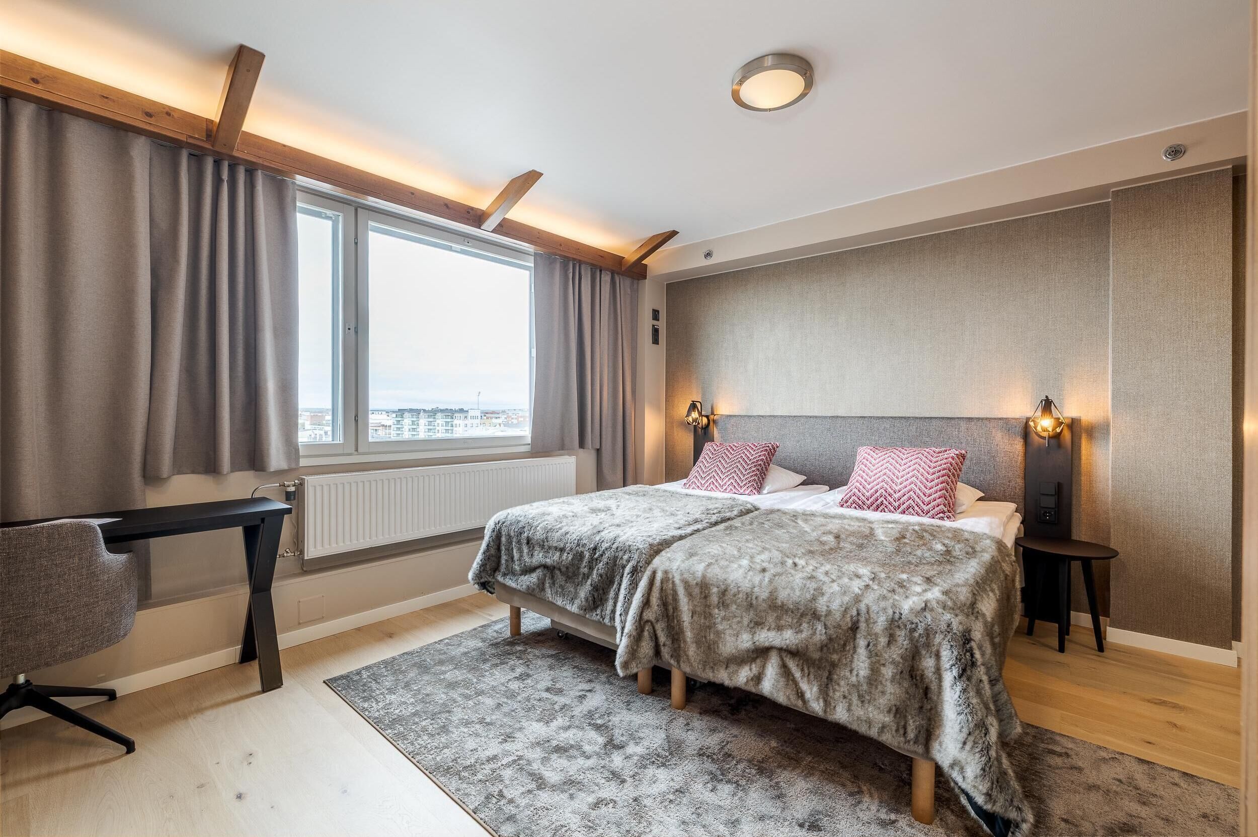 A bedroom with a bed, a desk, a chair, a window with curtains, and a city view.