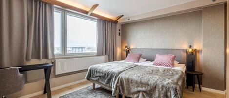 A bedroom with a bed, a desk, a chair, a window with curtains, and a city view.