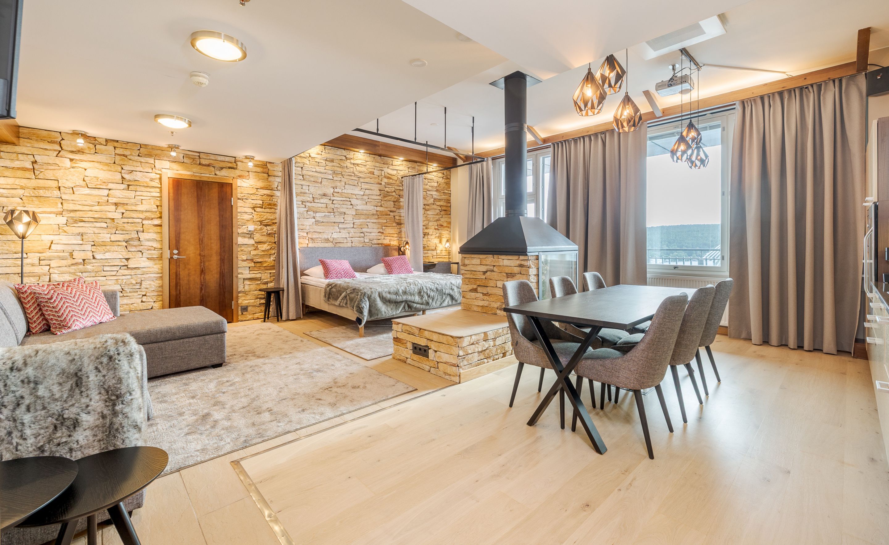 A modern living room with a stone wall, a fireplace, a dining area with chairs, and a large window with curtains.