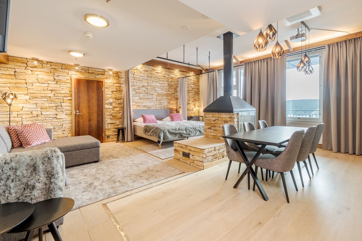 A modern living room with a stone wall, a fireplace, a dining area with chairs, and a large window with curtains.