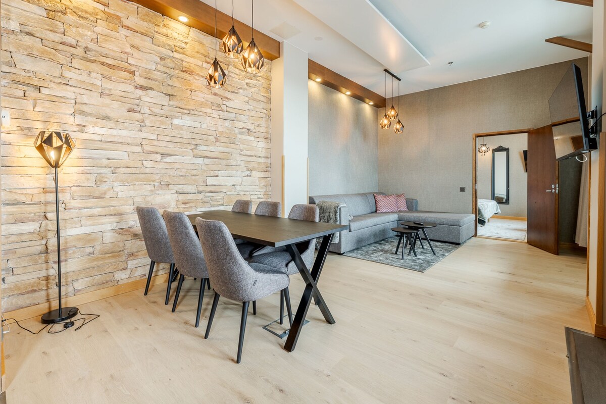 A modern living room with a stone wall, a wooden floor, a dining area with a table and chairs, and a sofa.