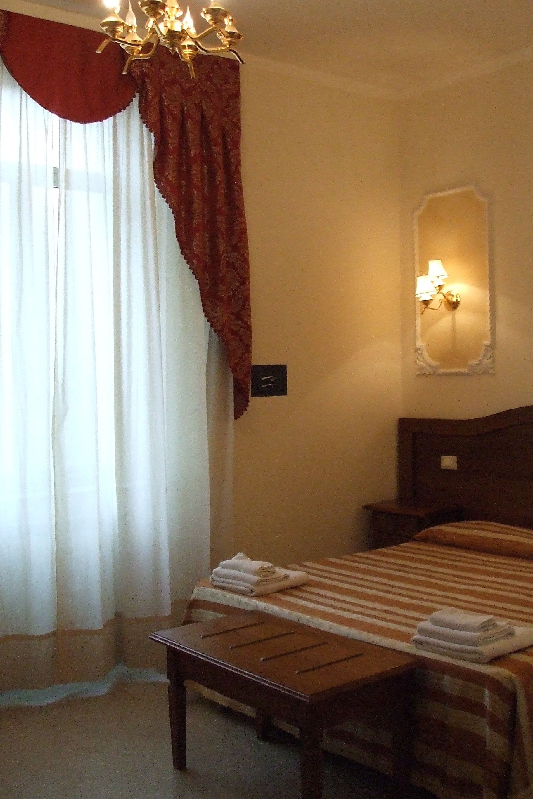 Camera d'albergo con un letto, un comodino, un tavolo, un lampadario, una finestra con tende e un punto luce a parete.