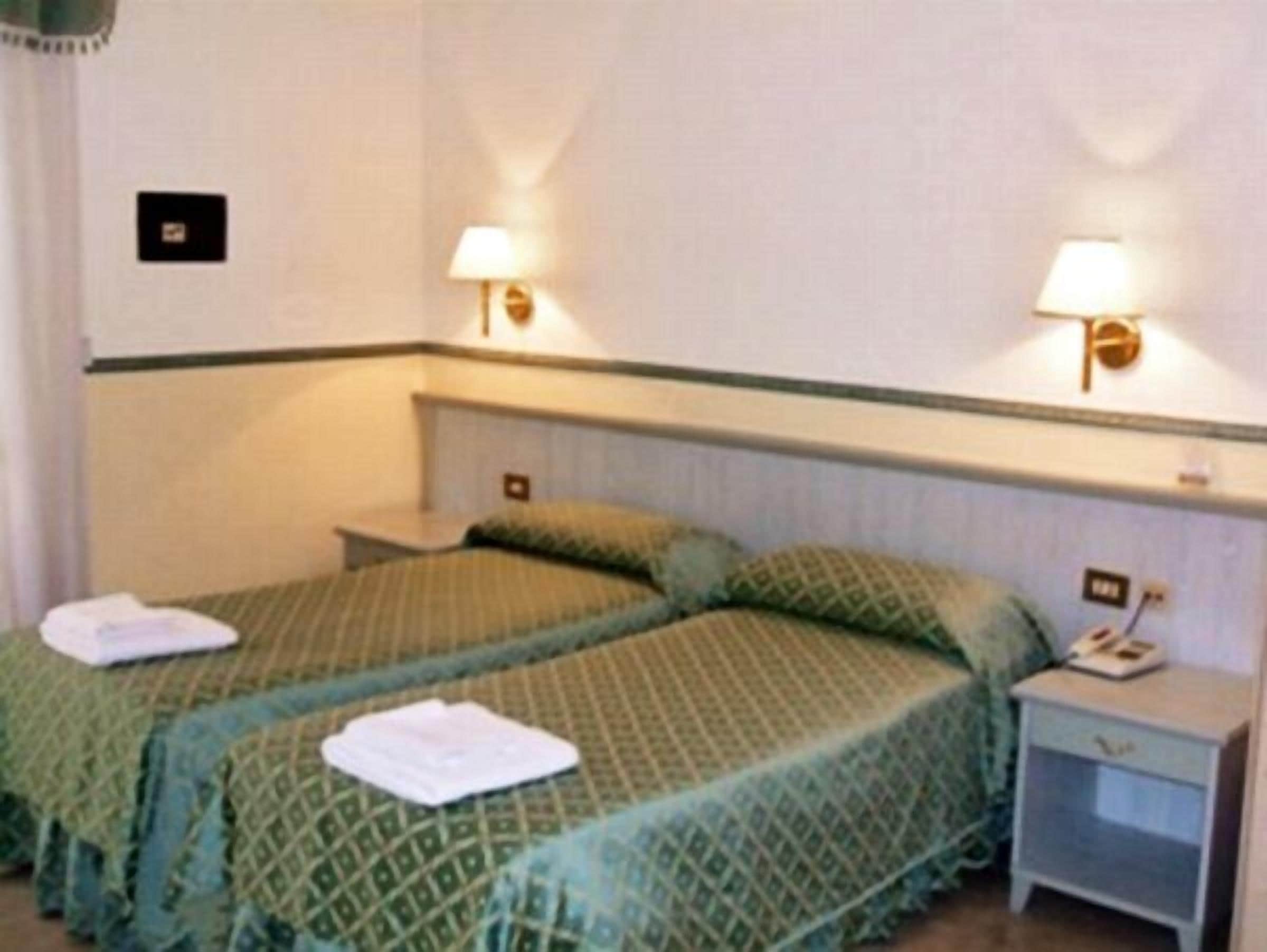 A hotel room with two beds, each with green bedding and pillows, a nightstand with a lamp, and a small table with a telephone.