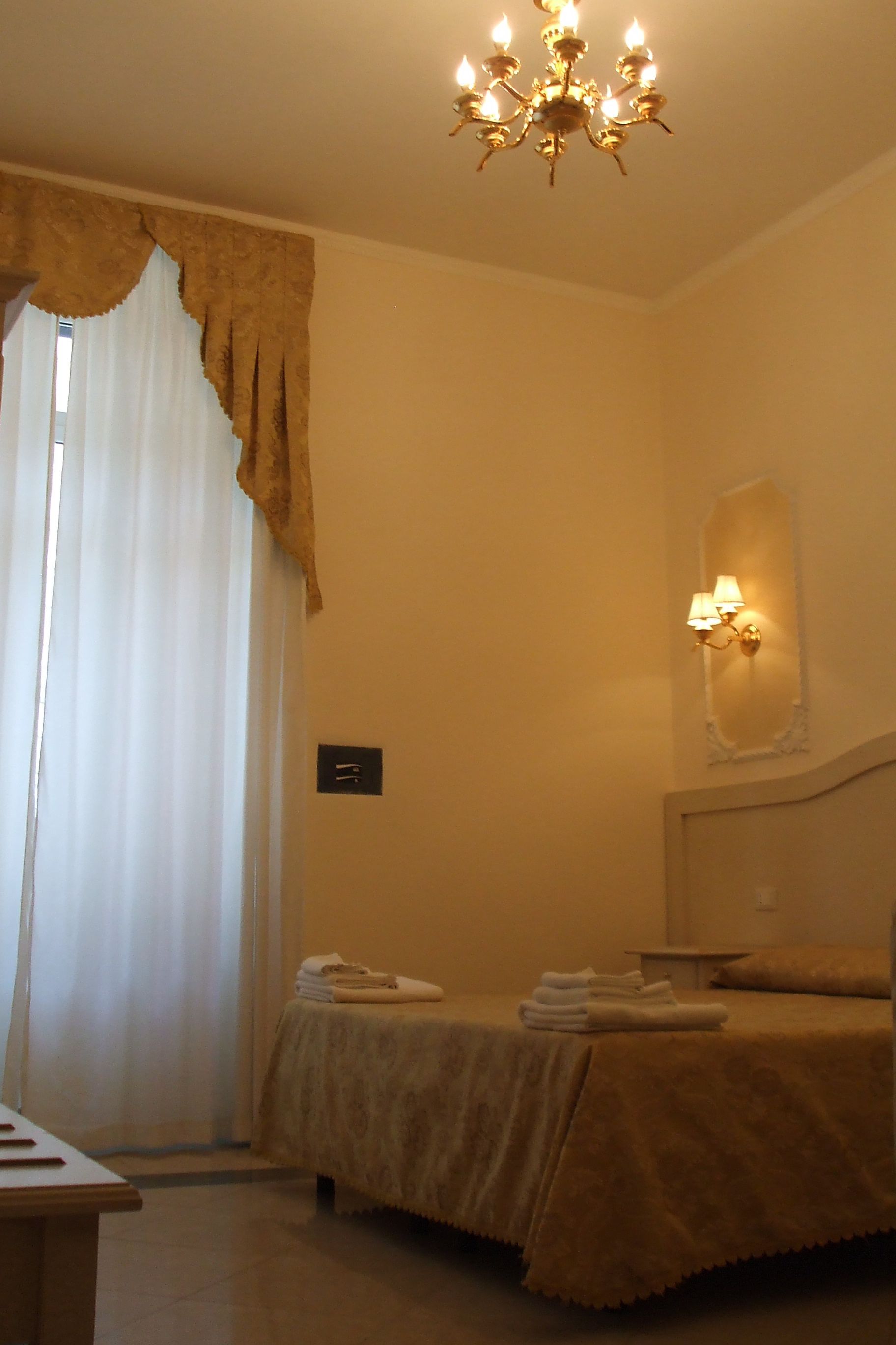 A hotel room with a bed, a chandelier, a wall-mounted lamp, and a window with curtains.