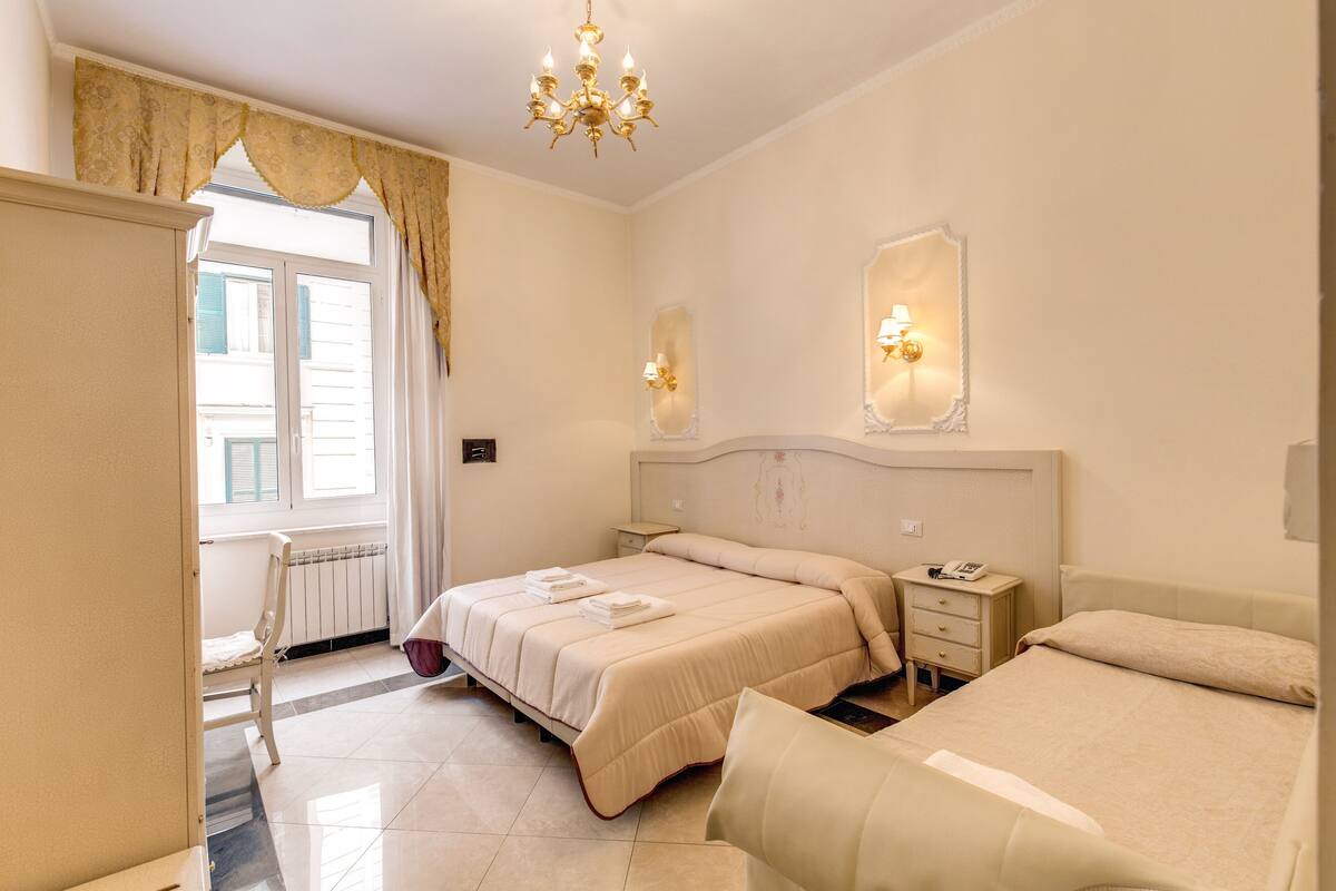 A hotel room with two beds, a chandelier, a window with curtains, and a bedside table with a lamp.