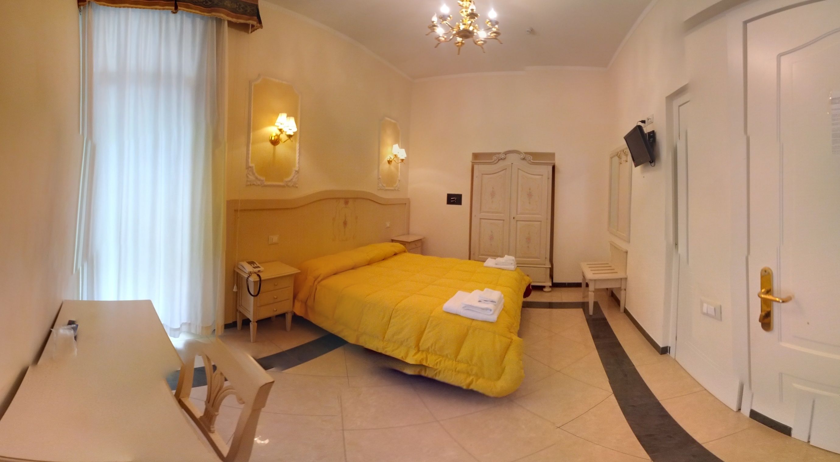 A hotel room with a bed, bedside tables, a desk, a wardrobe, and a chandelier.