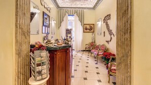 A hallway with a marble column, a reception desk with magazines, a chandelier, and a patterned floor. - Hotel Cambridge (Rome)