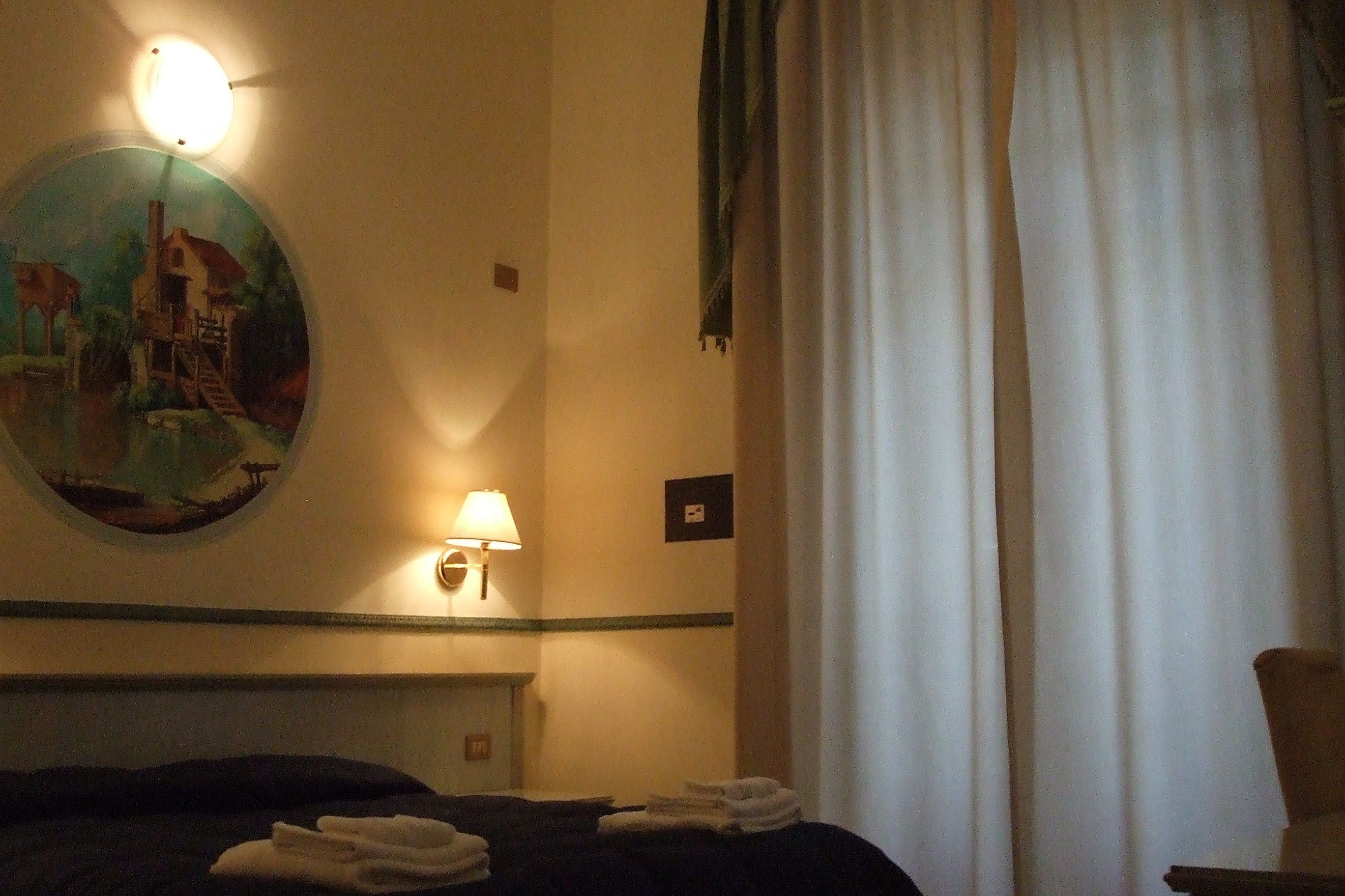 A hotel room with a bed, a wall-mounted lamp, a painting, and a window with curtains.