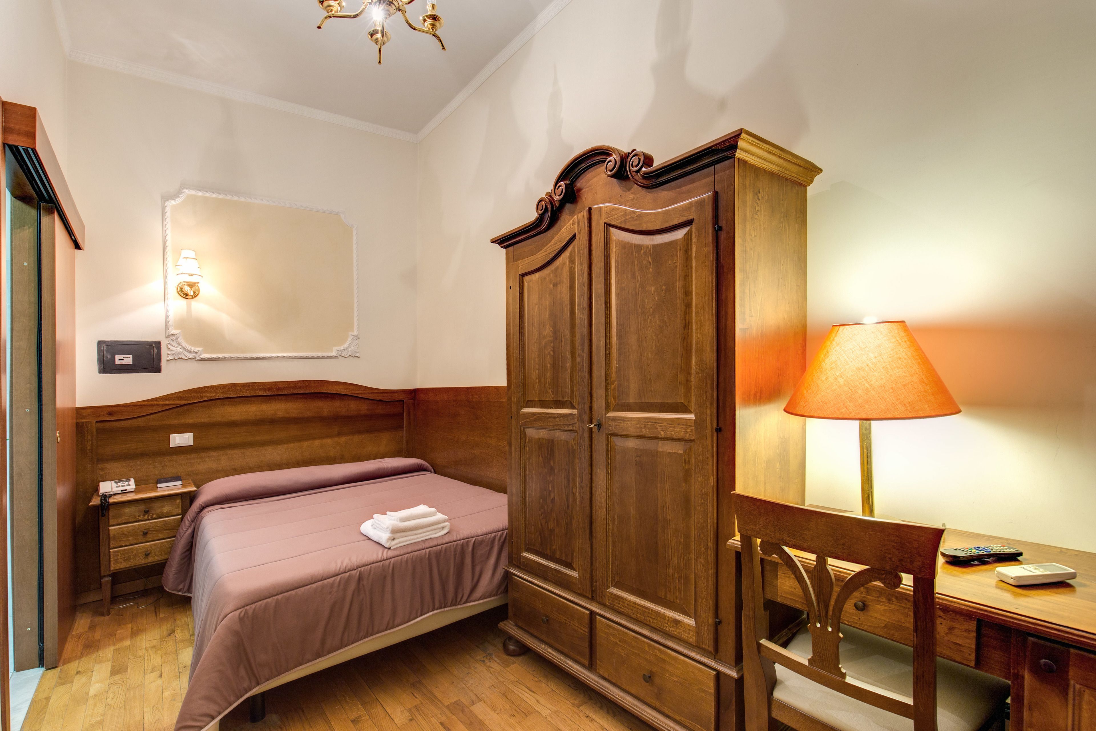 Un dormitorio con un armario de madera, una cama con edredón rosa, un escritorio con lámpara y una silla.