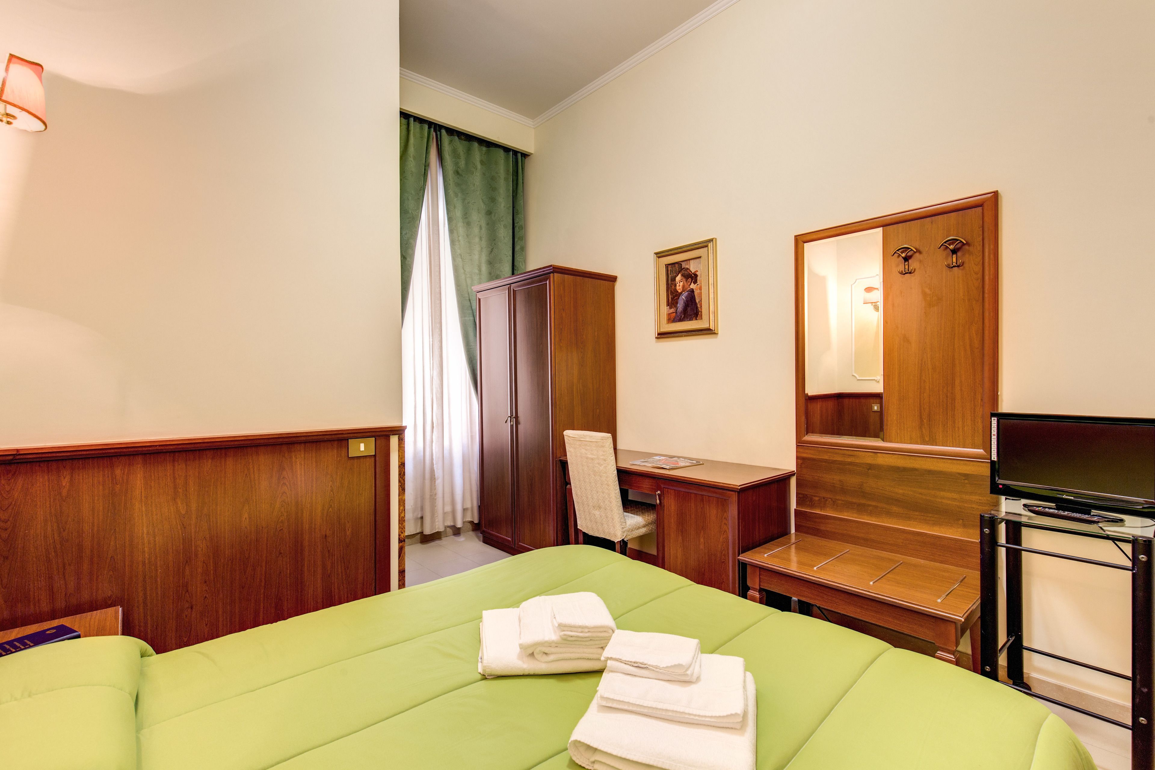 Ein Hotelzimmer mit einem grünen Bett, einem Schreibtisch mit Stuhl, einem Kleiderschrank, einem Fernseher und einem Bild an der Wand.