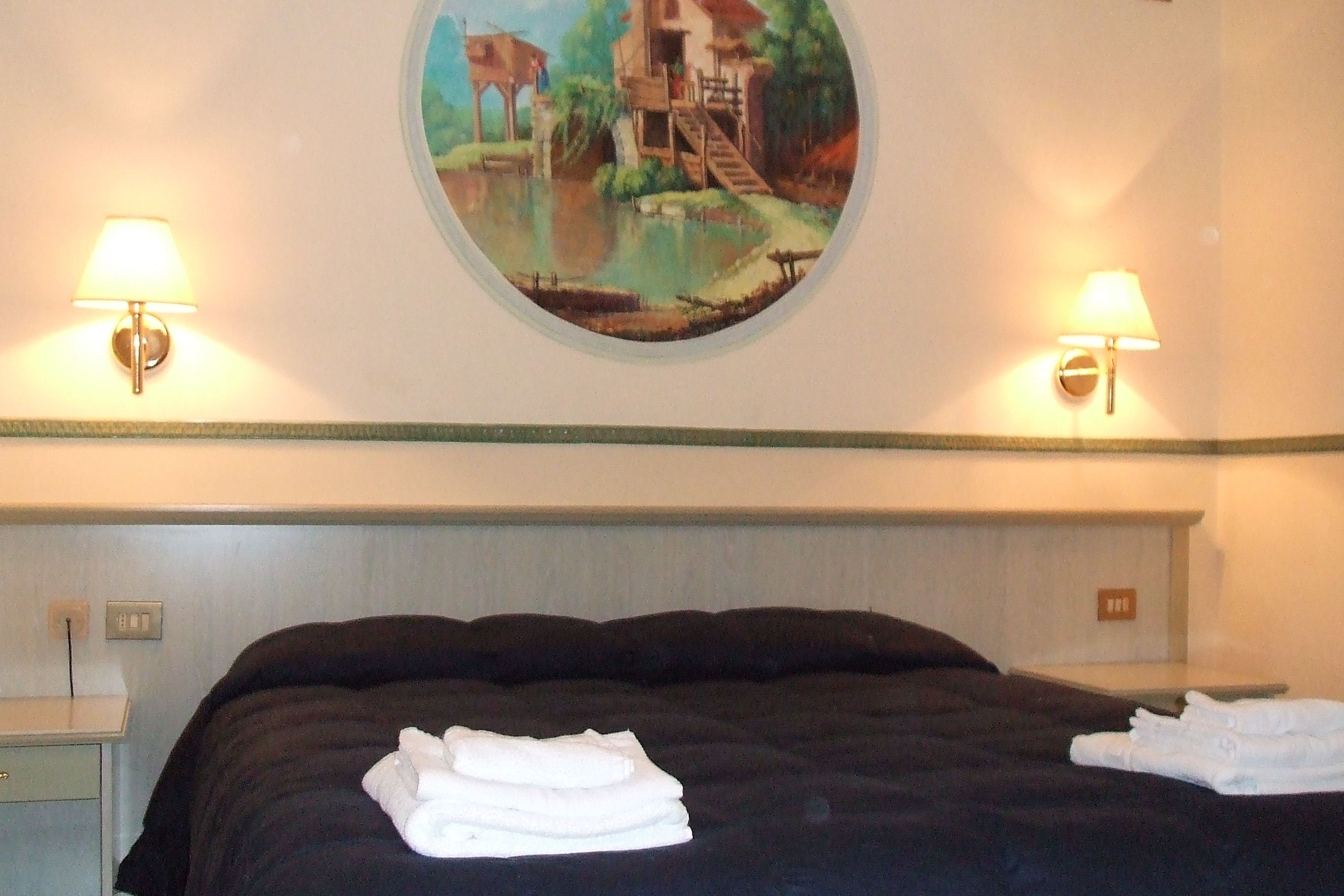 A hotel room with a bed, two wall-mounted lamps, a painting of a house by a lake, and a nightstand with a lamp.
