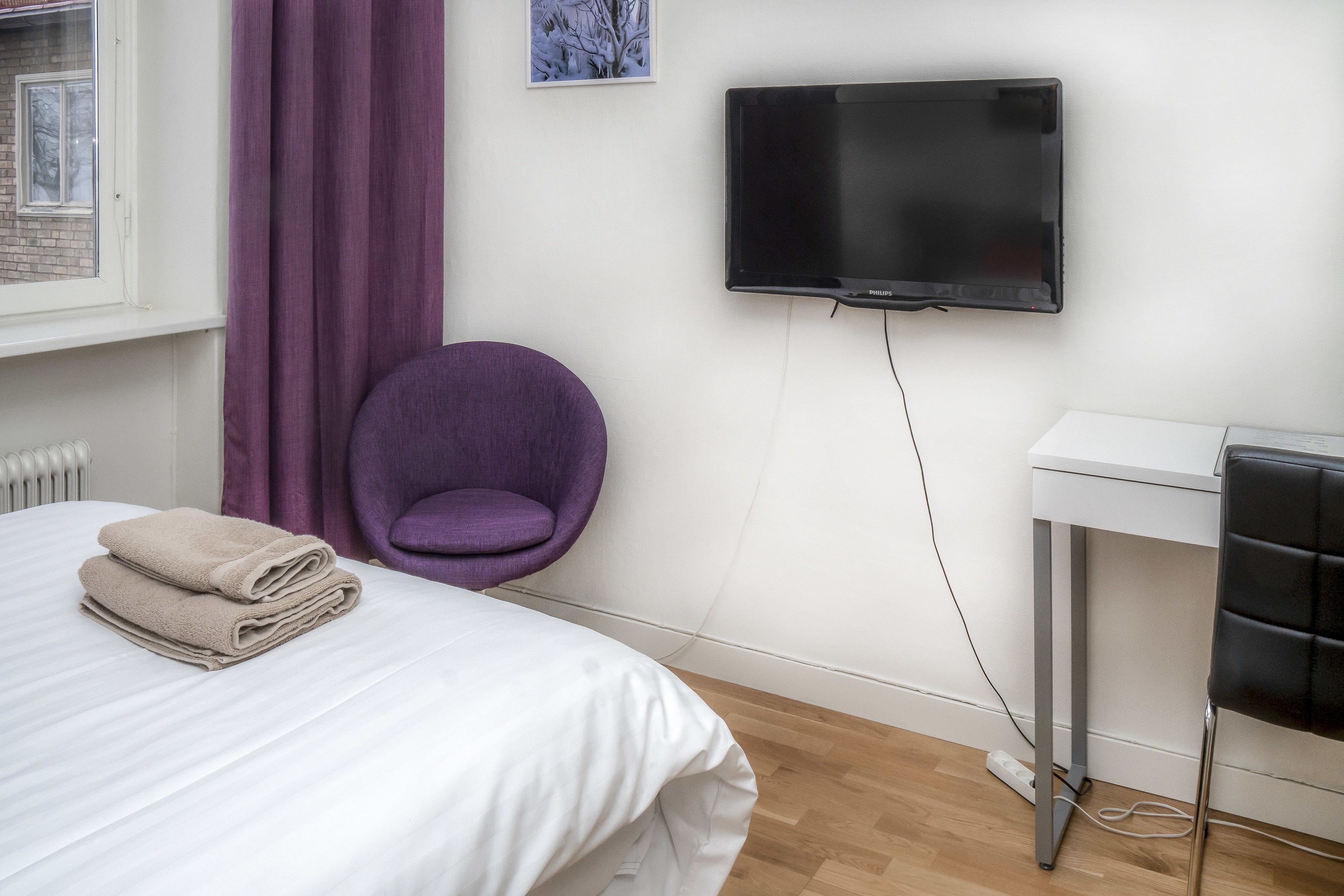 A neatly made bed with folded towels, a purple chair, a television mounted on the wall, and a white table with a black chair.