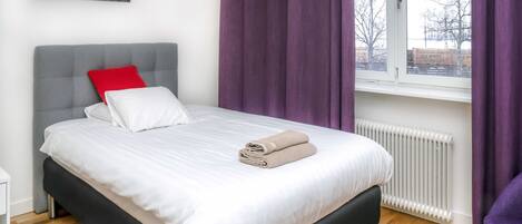 Una cama bien hecha con almohadas blancas y rojas, una toalla doblada y un cabecero gris.
