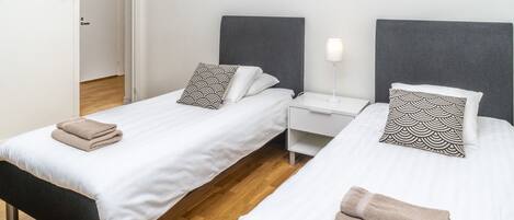 A hotel room with two beds, a wooden floor, a white wall, a painting of a sunset over water, and a door with a calendar and a map.