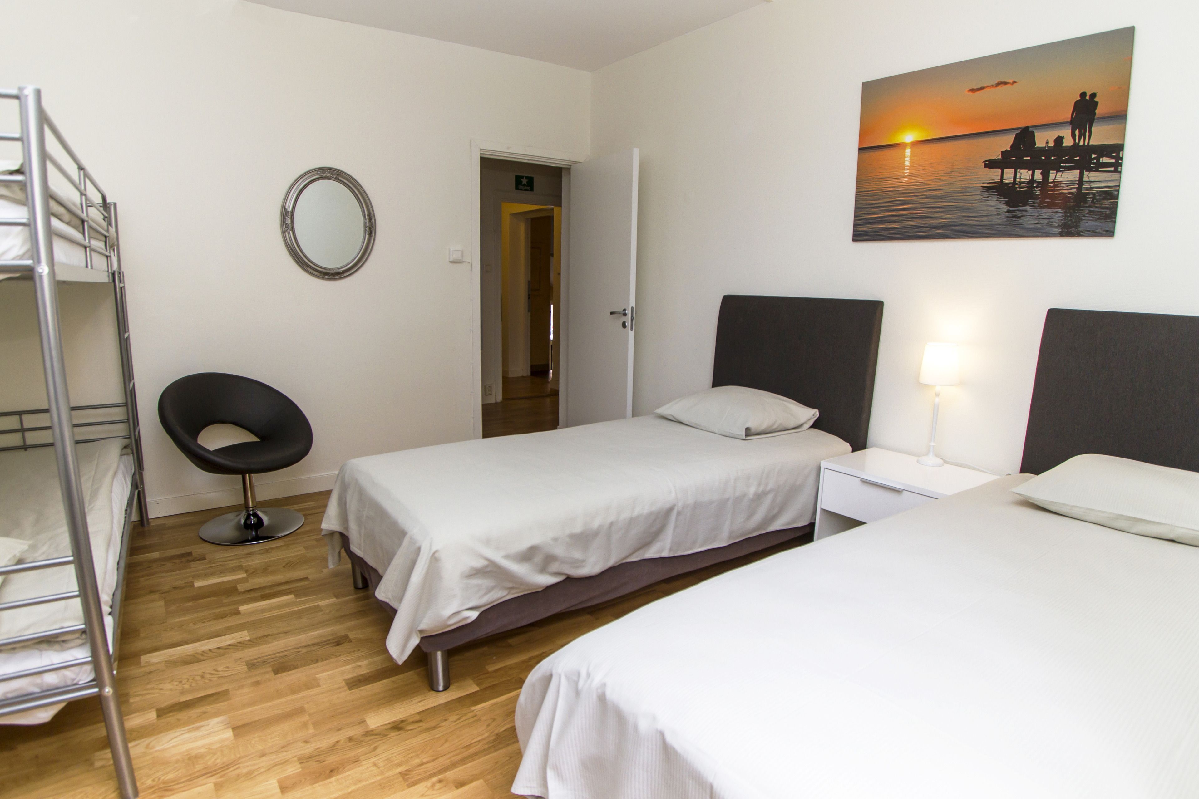 A hotel room with two single beds, a round mirror, a wooden floor, a door leading to another room, and a painting of a sunset over water.