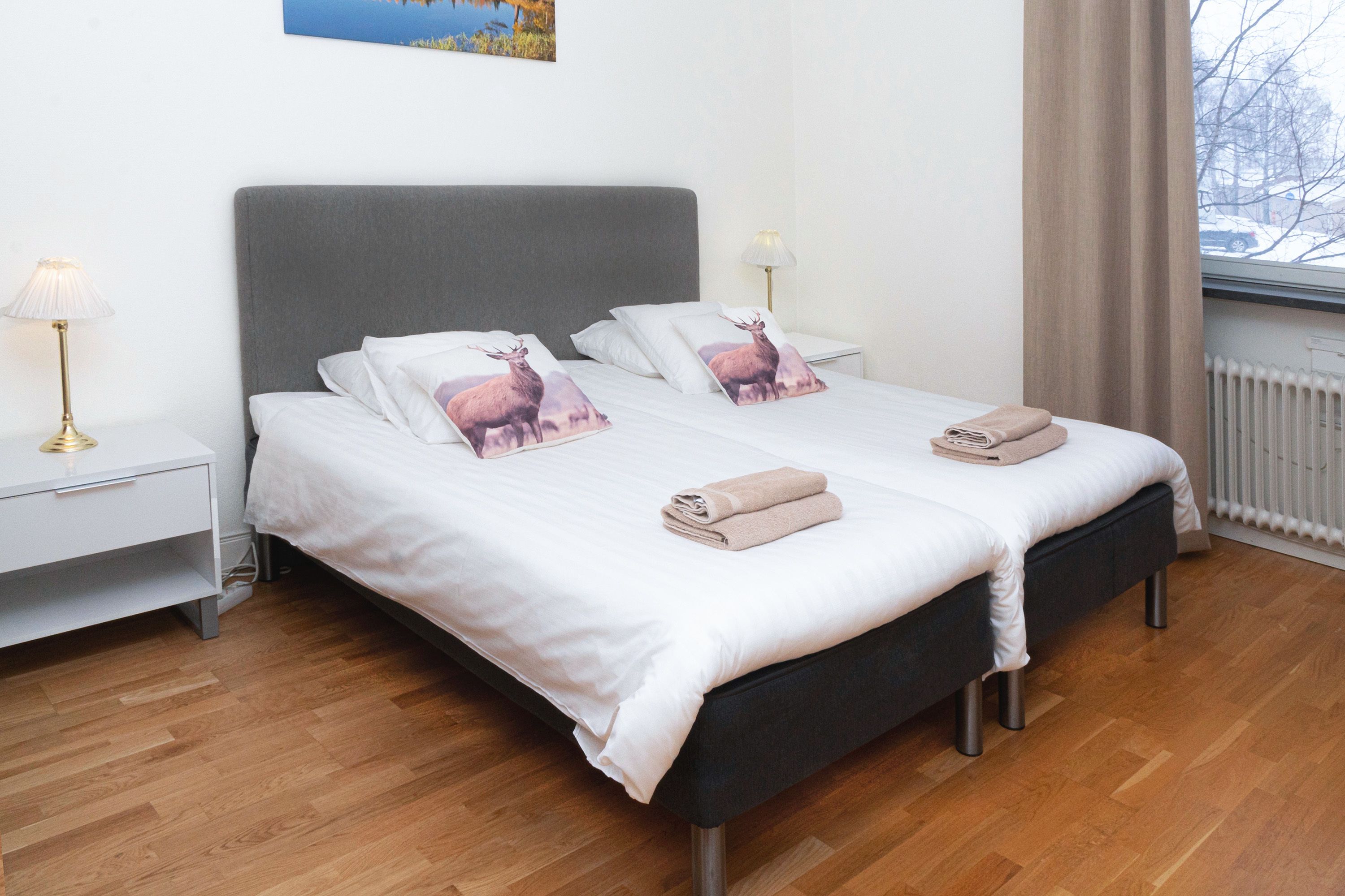 A neatly made double bed with white linens and two pillows featuring a deer design, a bedside table with a lamp, and a window with a view of a snowy landscape.