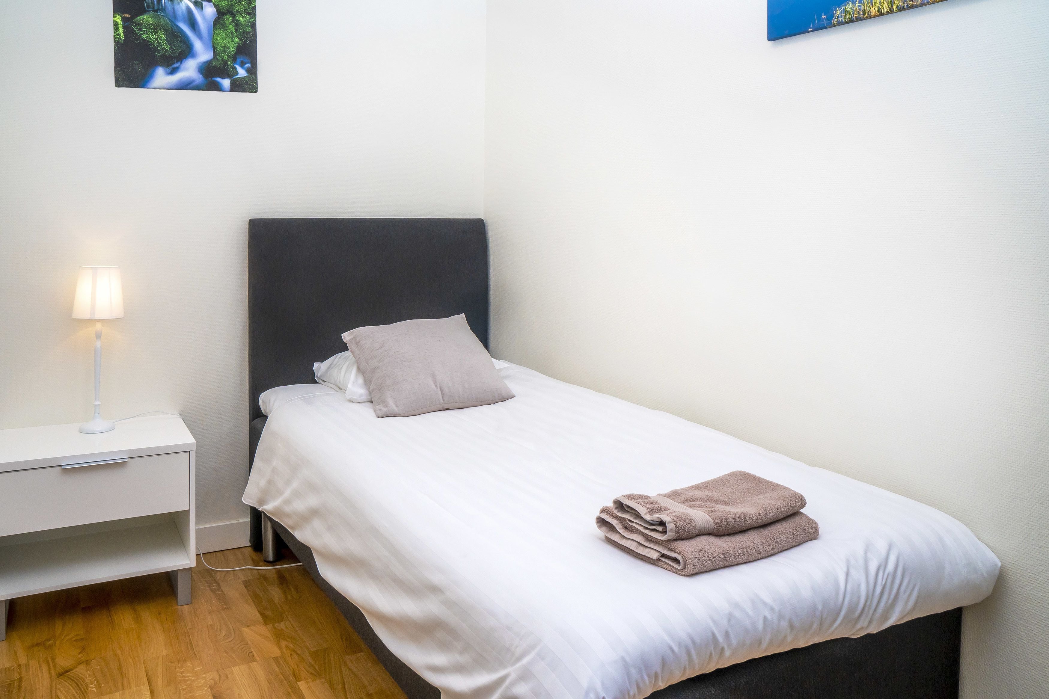 A neatly made bed with a pillow and a folded towel, a bedside lamp, and a small nightstand.