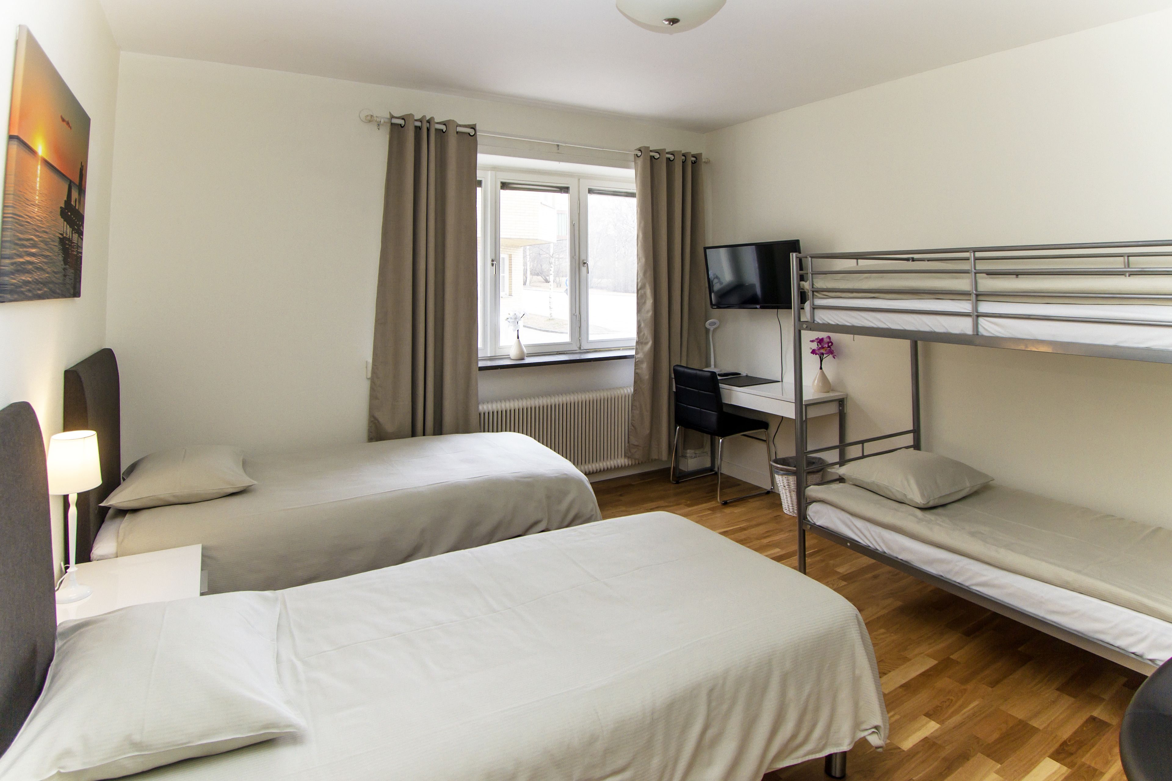 Ein Zimmer mit zwei Einzelbetten, einem Schreibtisch mit Stuhl, einem Fernseher und einem Fenster mit Vorhängen.