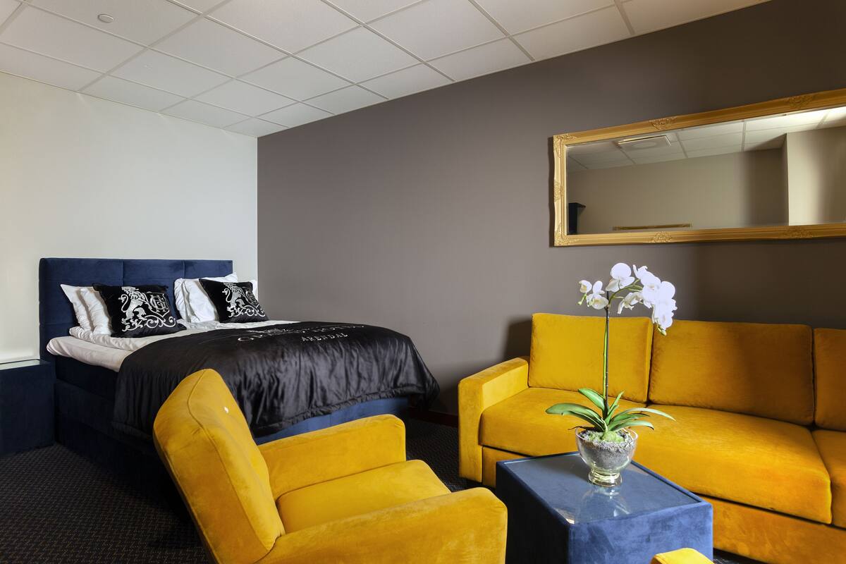 Una habitación de hotel con una cama, un sillón amarillo, un sofá y una pequeña mesa con un florero.