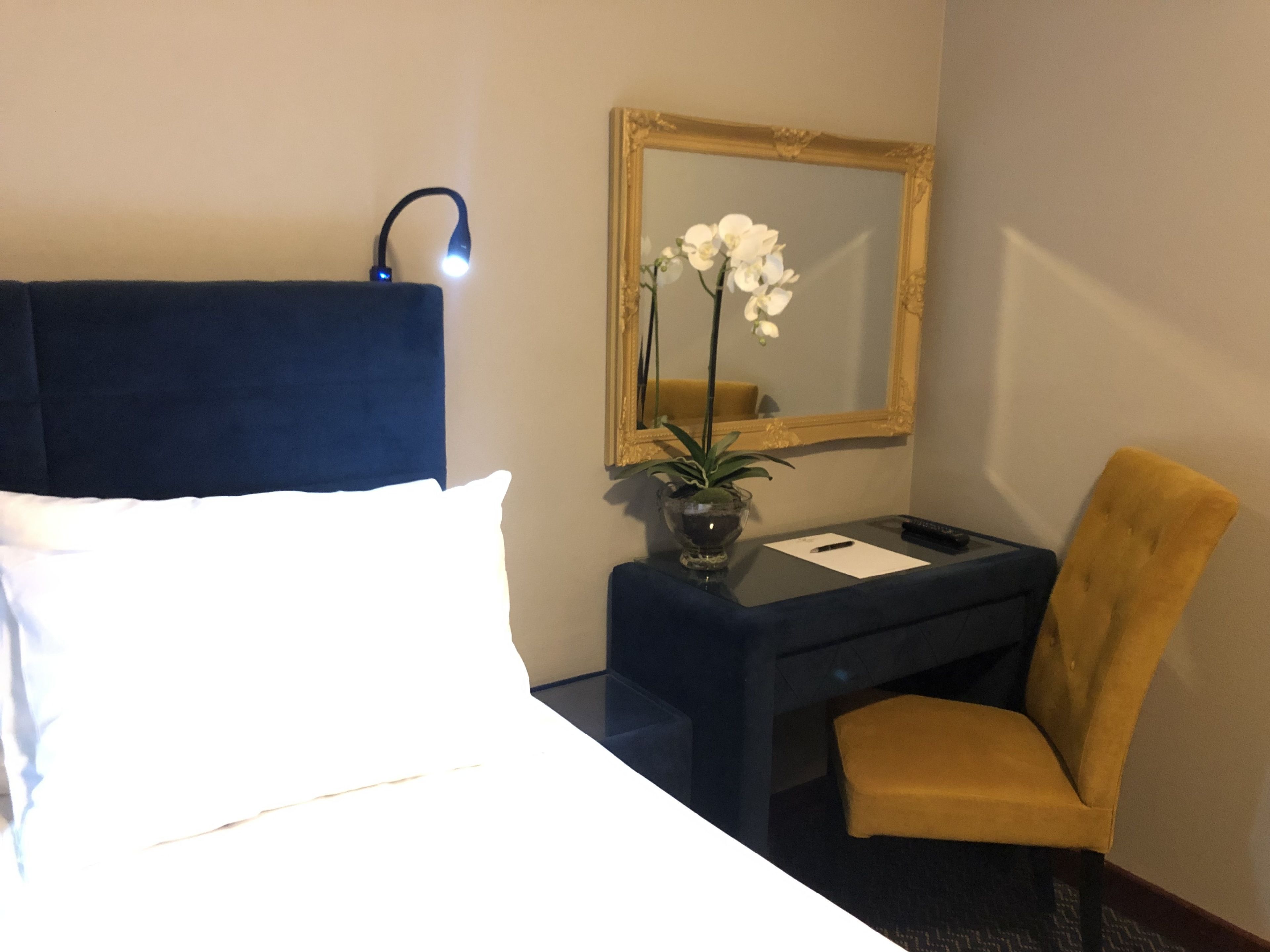 A hotel room with a bed, a desk, a chair, a mirror, and a vase with flowers.