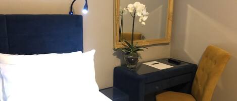 A hotel room with a bed, a desk, a chair, a mirror, and a vase with flowers.