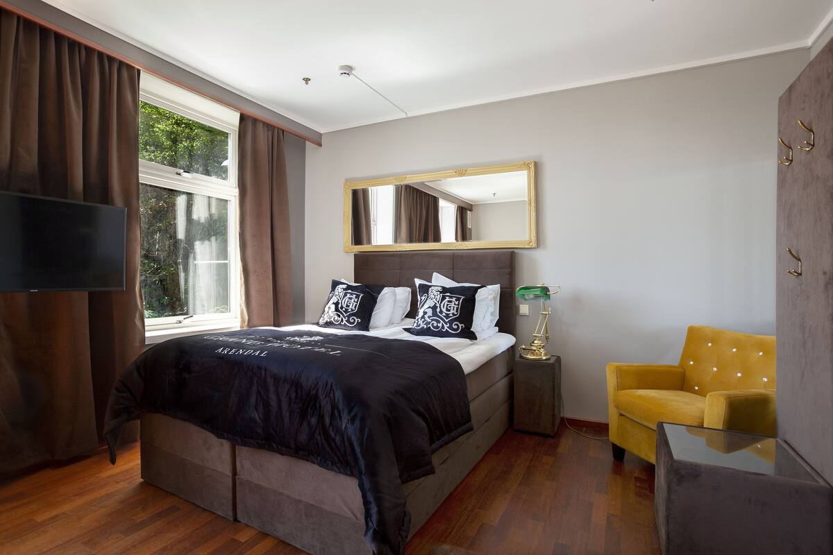 Un dormitorio con cama, televisor, espejo, sillón amarillo y una pequeña mesa.