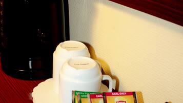 A coffee station with a coffee maker, two white cups, and a basket containing magazines and a pen.