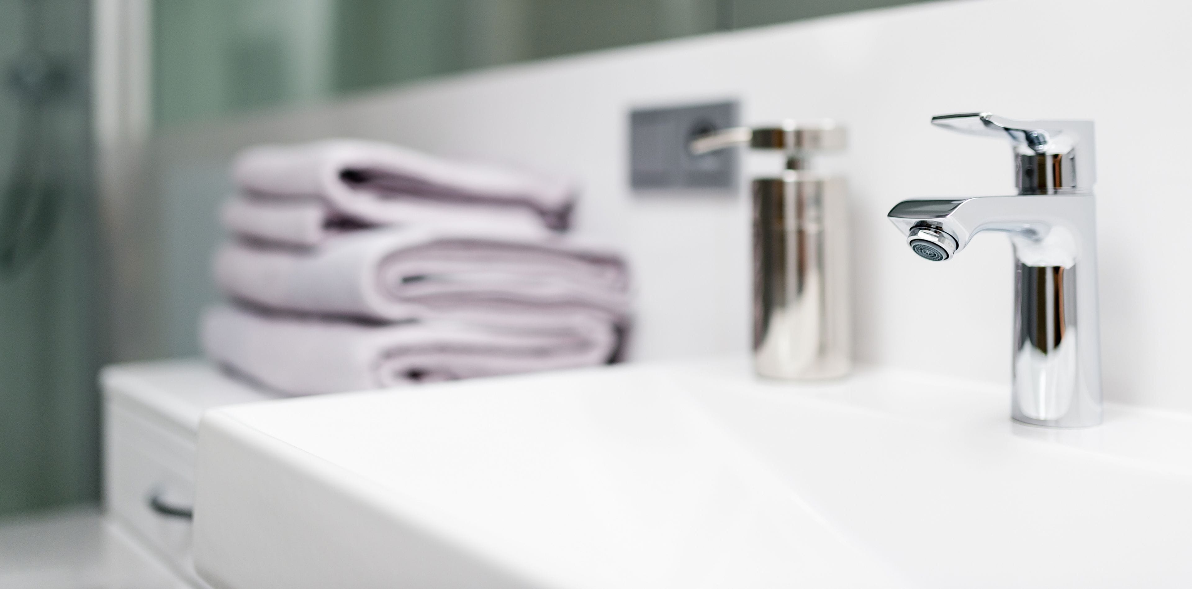 A bathroom with a white sink, two chrome faucets, and a stack of folded towels.