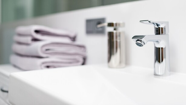 Un baño con un lavamanos blanco, dos grifos cromados y una pila de toallas dobladas.