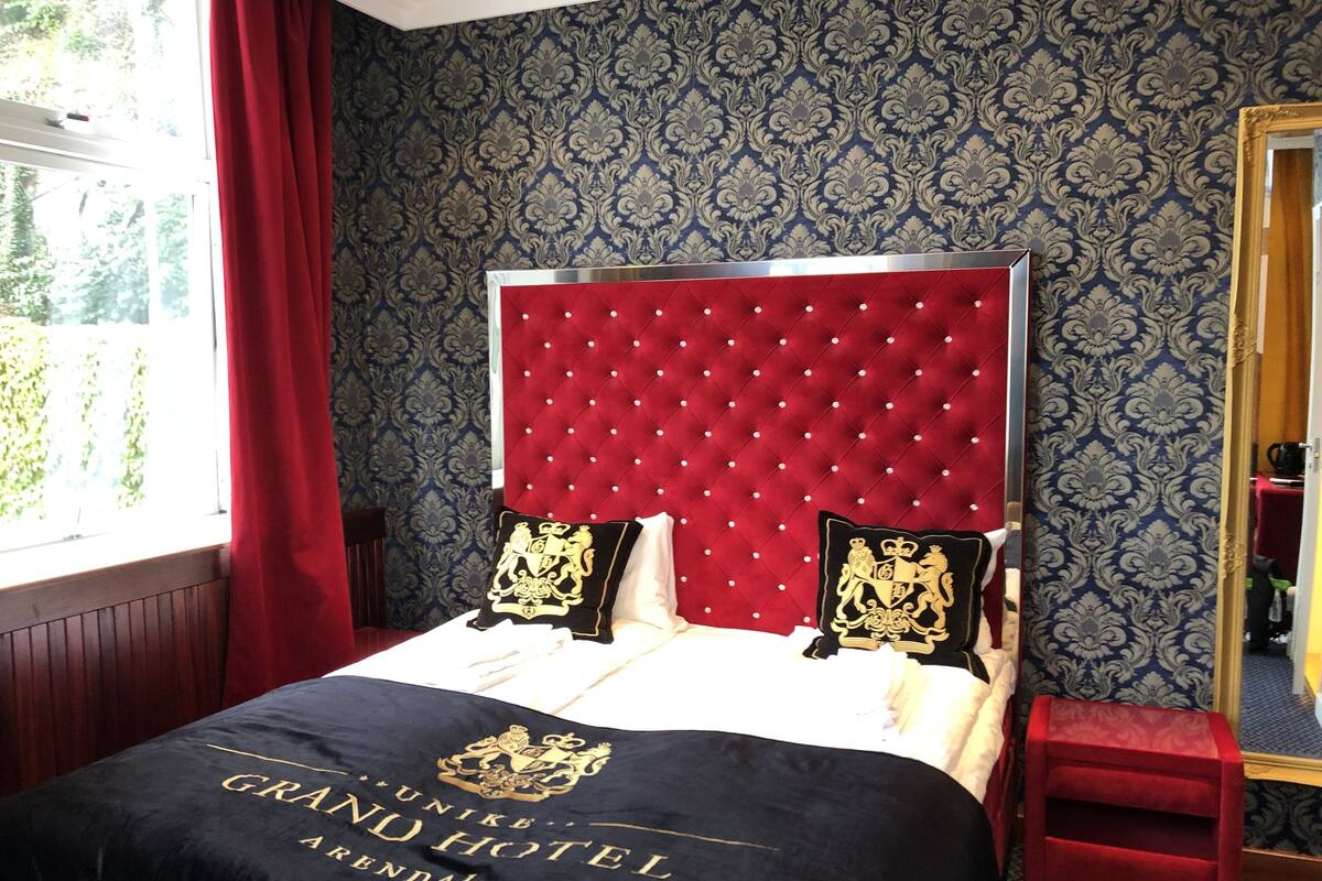 Habitación de hotel con cabecera tapizada roja, papel pintado estampado y cama con colcha negra que presenta un escudo.