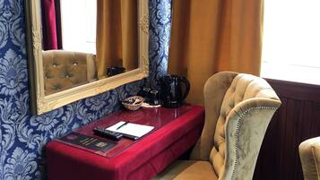 A room with a red desk, a beige armchair, a mirror, and a patterned wallpaper.