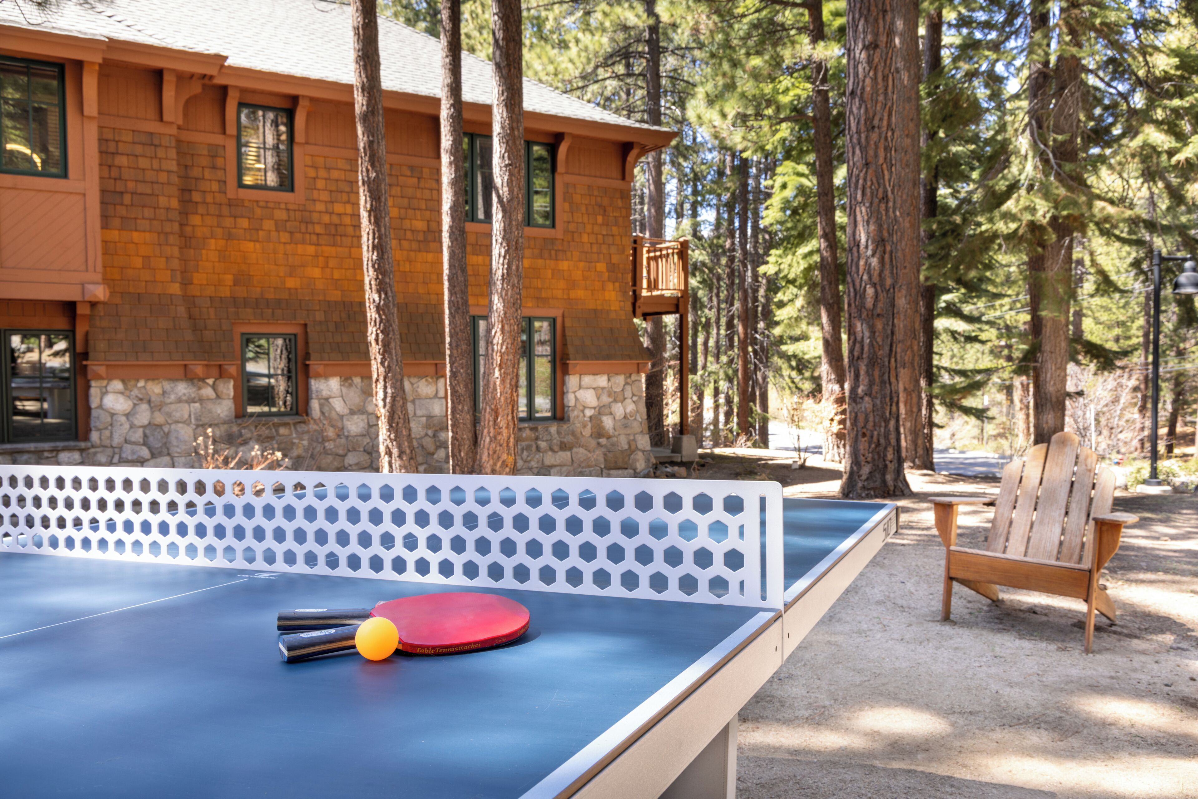A ping pong table with balls, a wooden chair, and a building surrounded by trees.