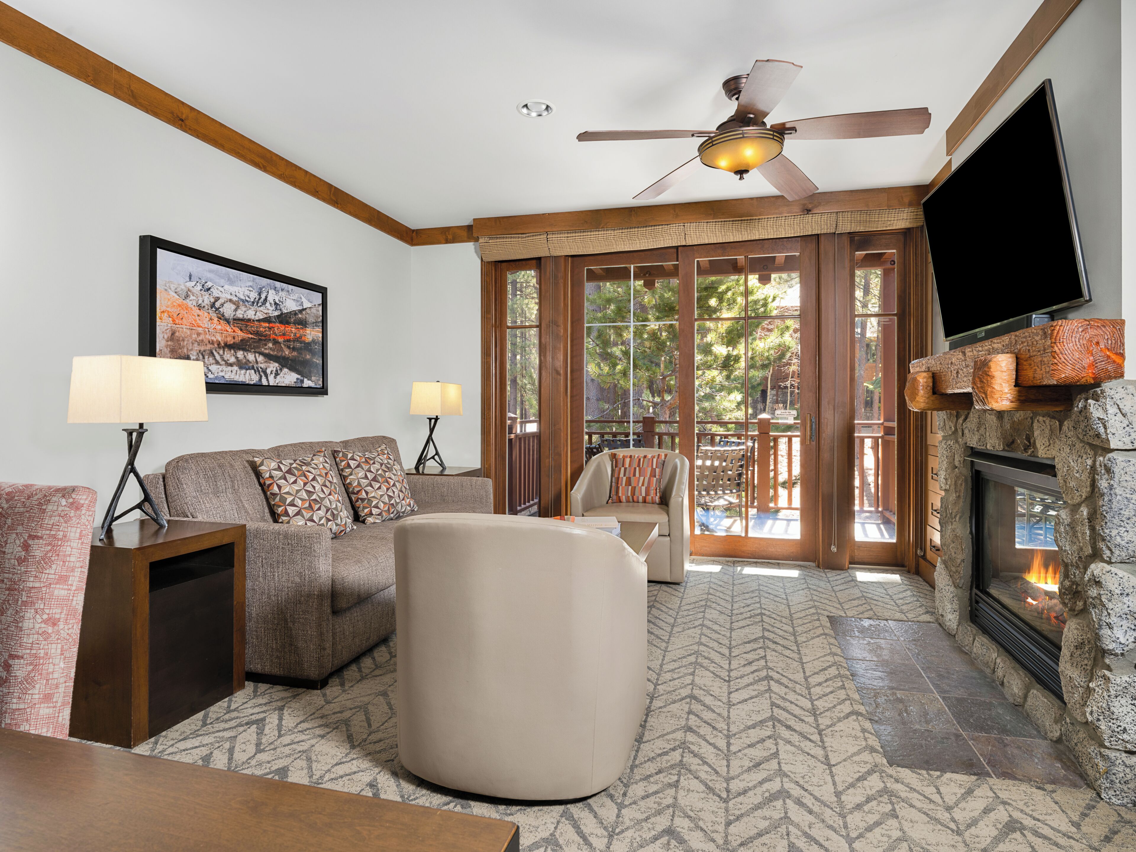A living room with a fireplace, a ceiling fan, and a large window with a view of trees.