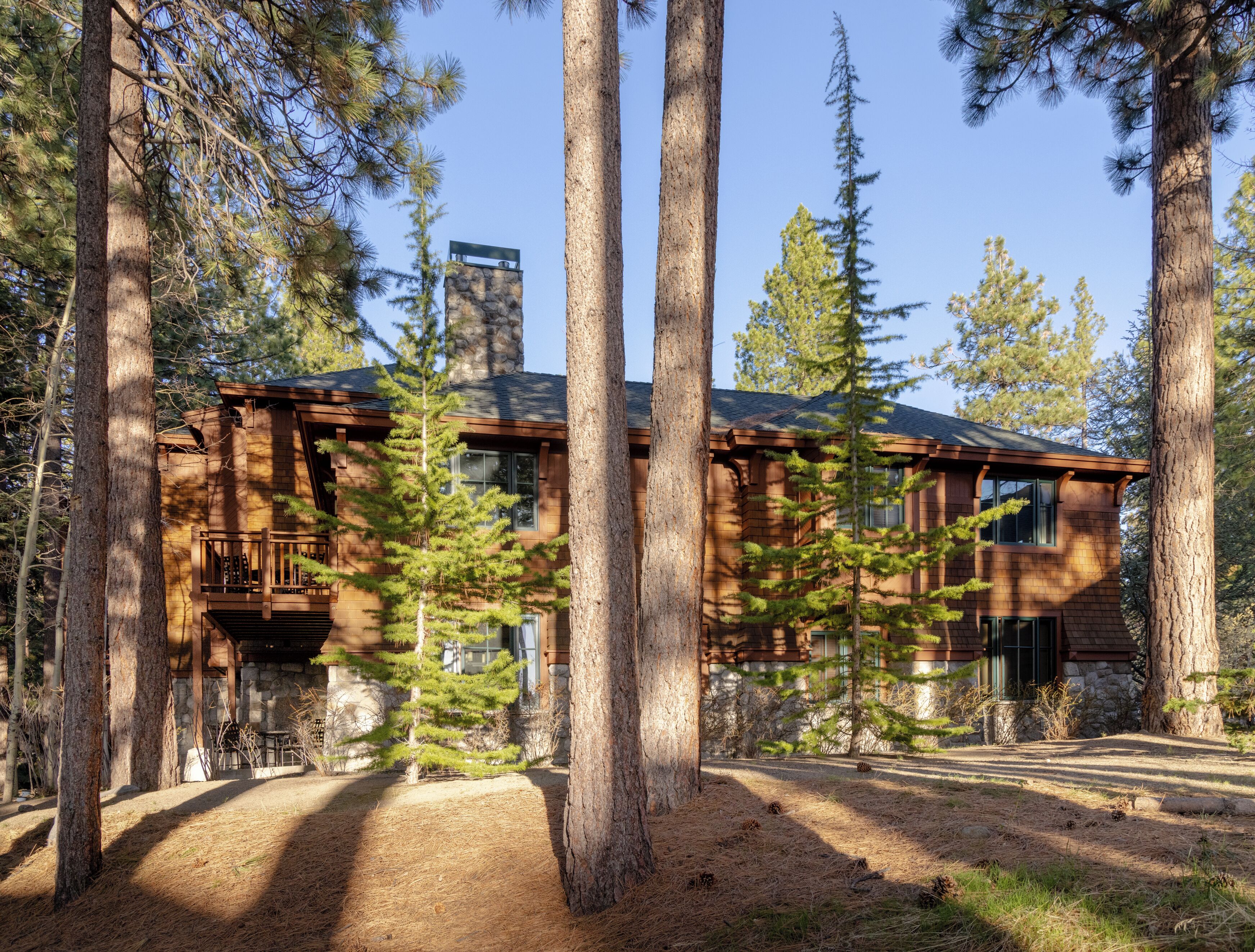A two-story log cabin with a stone foundation, surrounded by tall pine trees.
