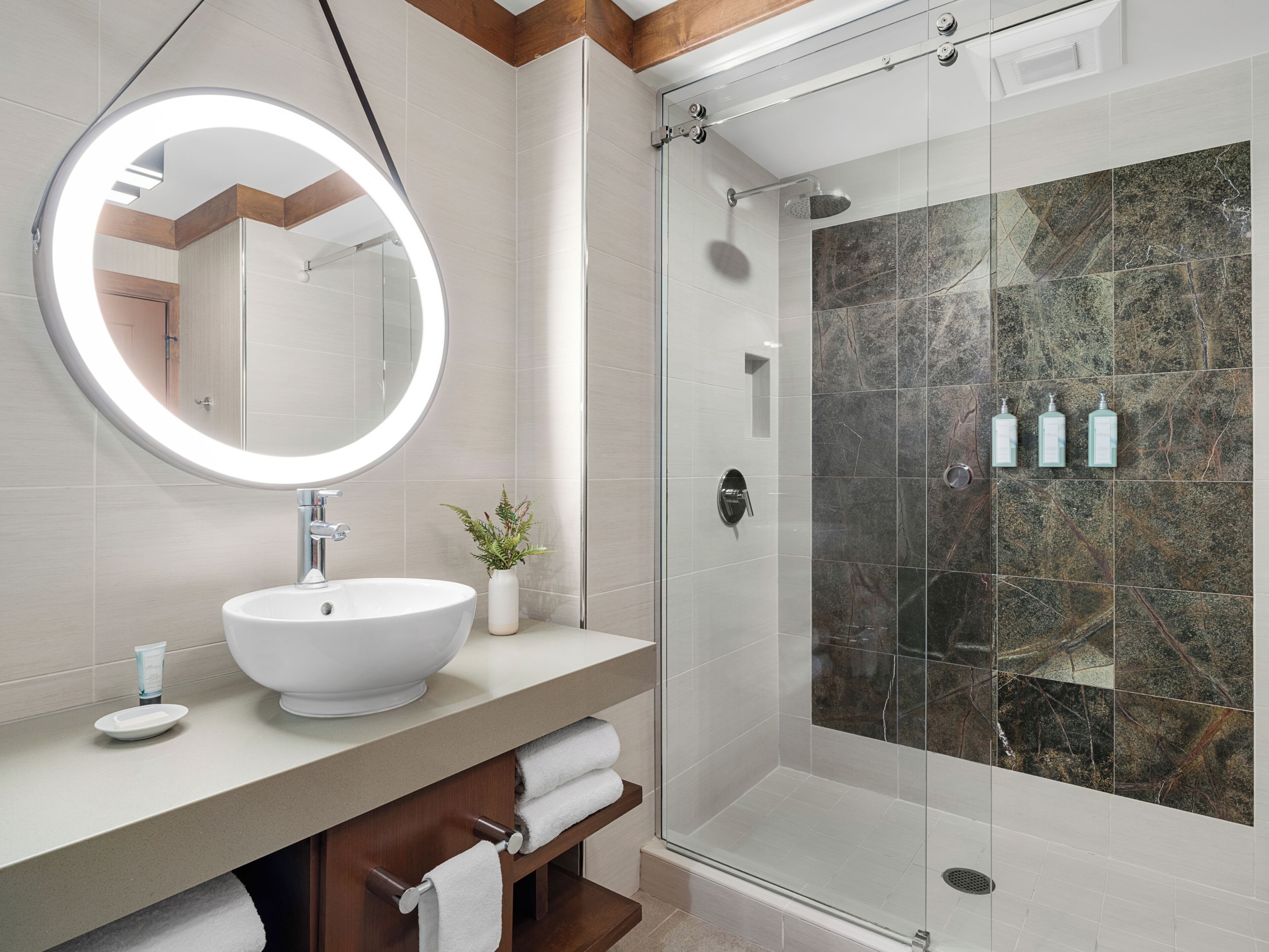 Un baño moderno con un espejo redondo, un lavamanos grande, una ducha con mampara de vidrio y una planta pequeña en el mueble de tocador.
