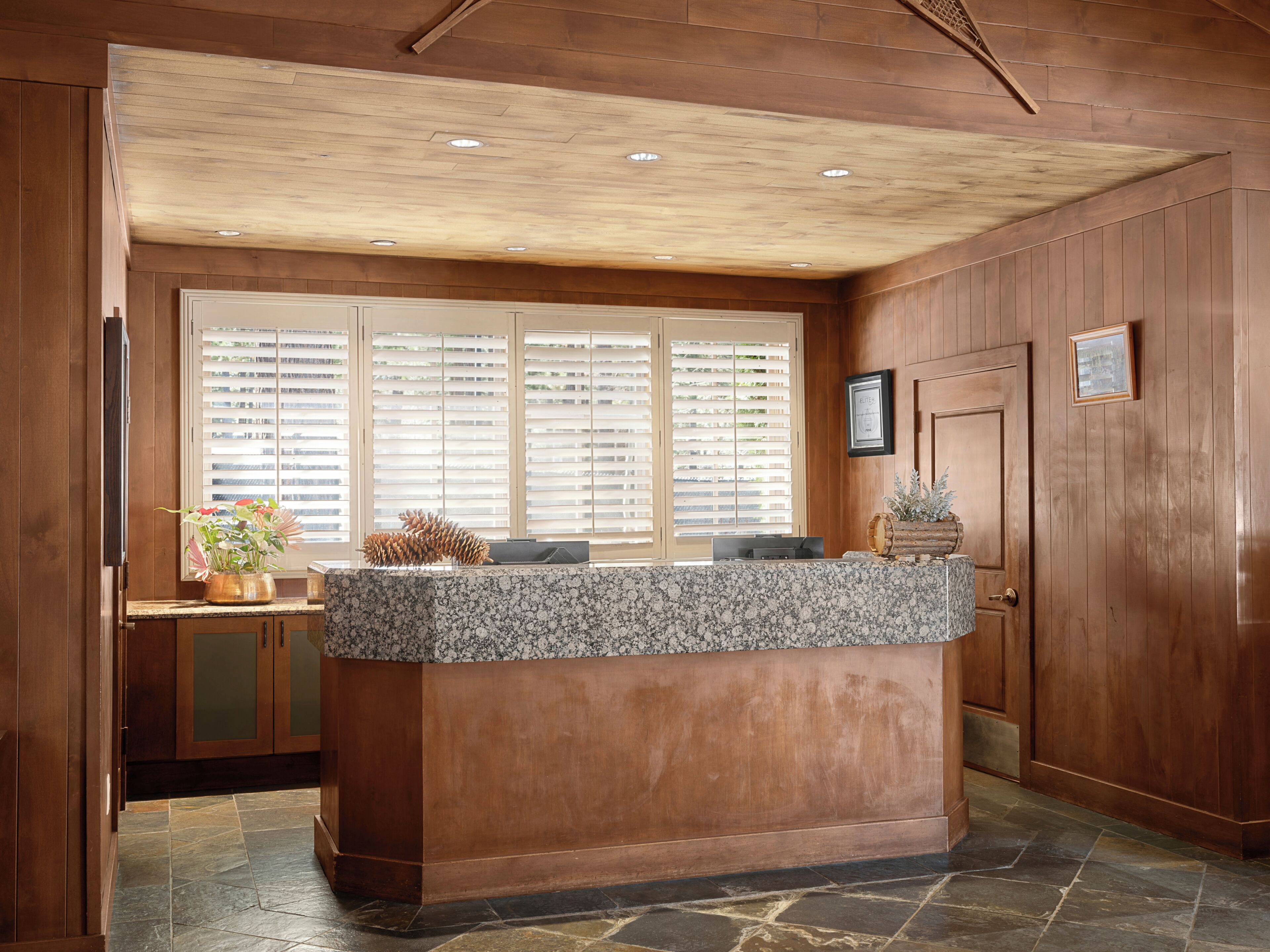 A kitchen with wooden paneling, a granite countertop, and a window with blinds.