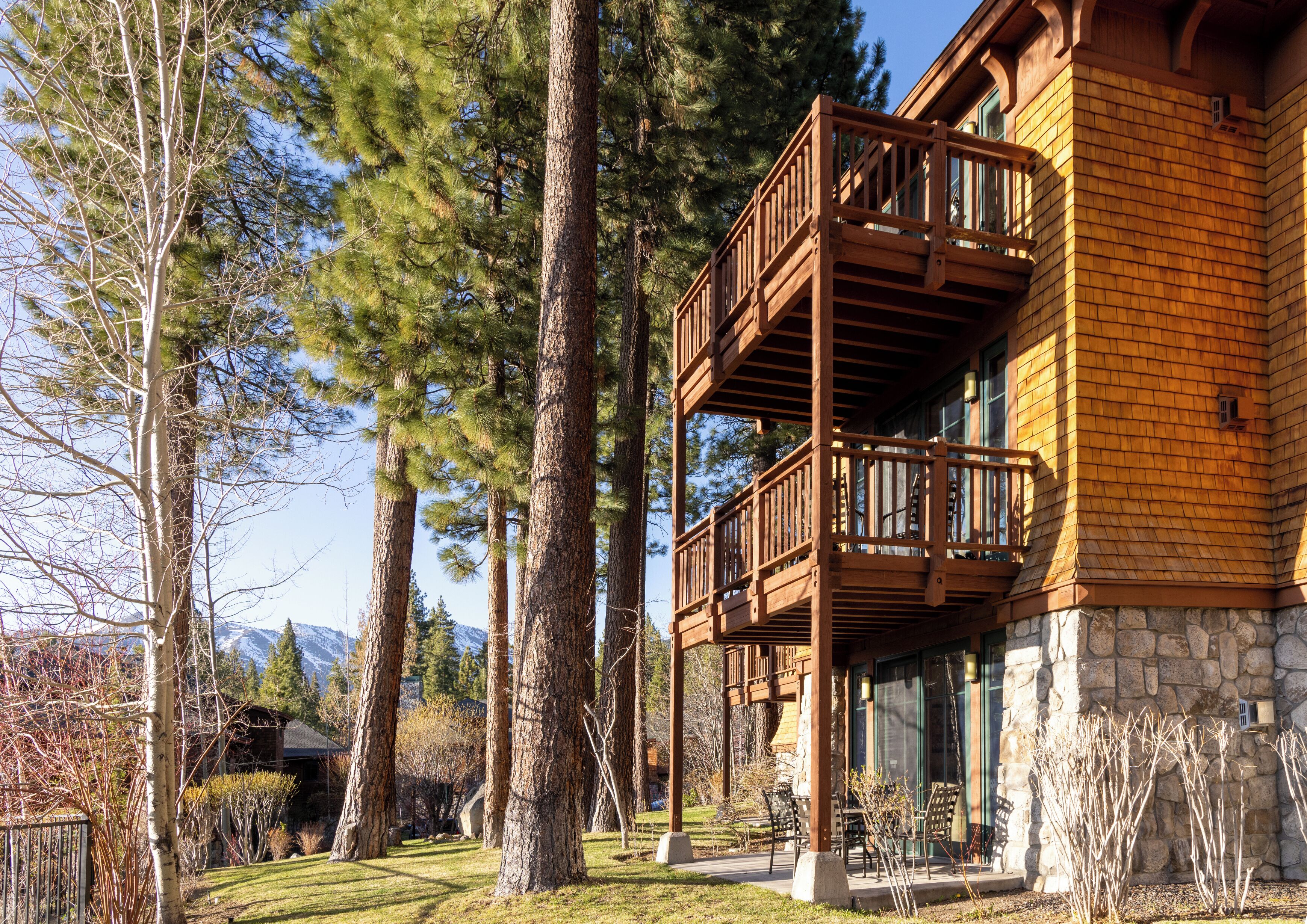 A two-story wooden cabin with a balcony, surrounded by tall trees and a mountain backdrop.