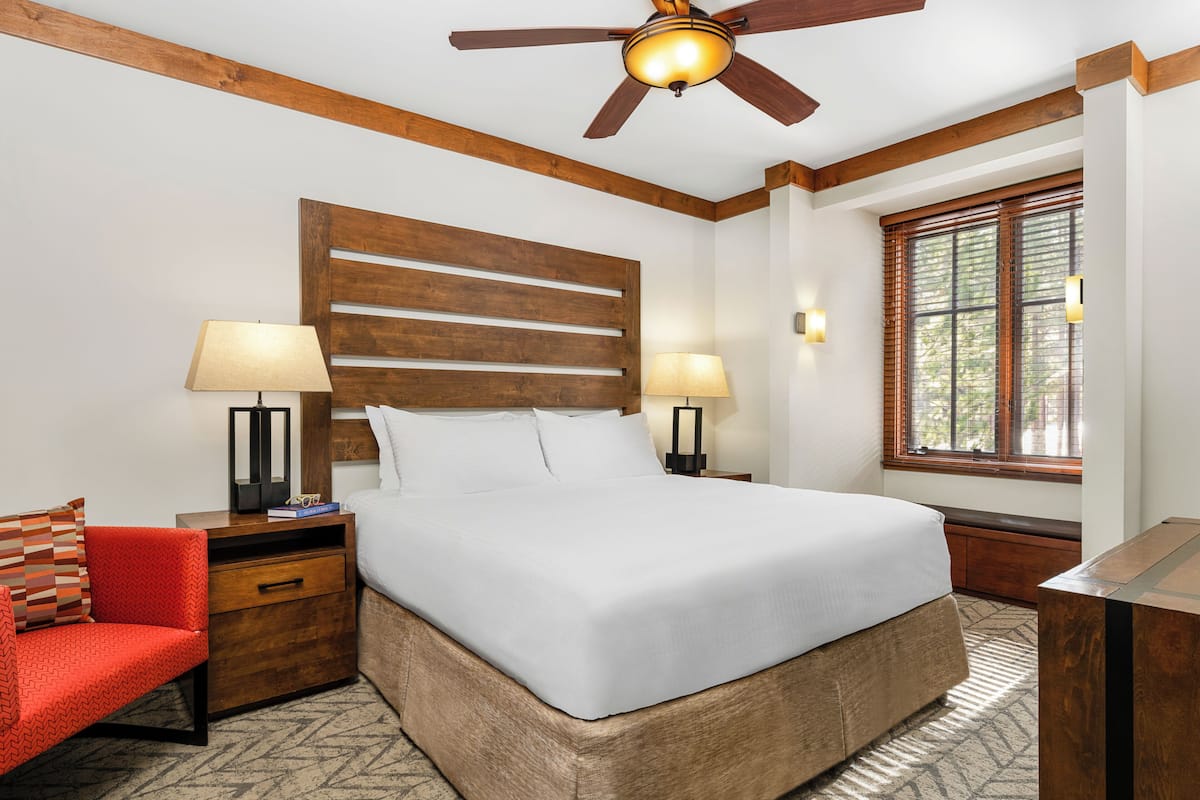 Habitación de hotel con una cama grande, cabecera de madera, dos lámparas de noche, una silla roja, una mesita de noche de madera y un ventilador de techo.