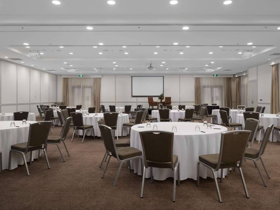Sala de conferências com mesas redondas, cadeiras, tela de projetor e quadro branco.