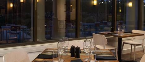A dining table set for two with clear glassware and cutlery, positioned in front of large windows overlooking an outdoor seating area.